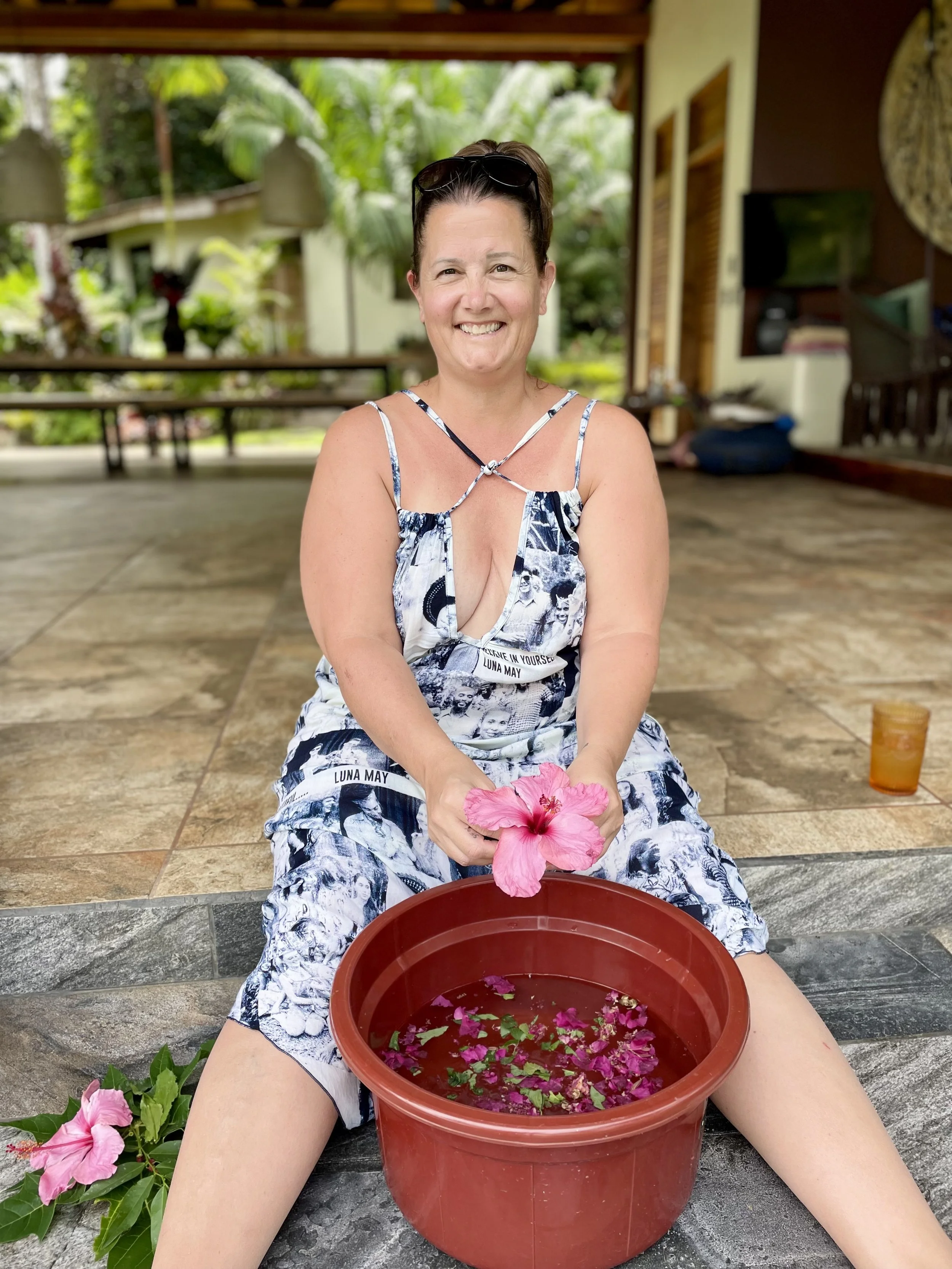 Woman sat on floor holding a pink flower, red container filled with water and other plants, floral bath preparation for Ayahuasca ceremony 