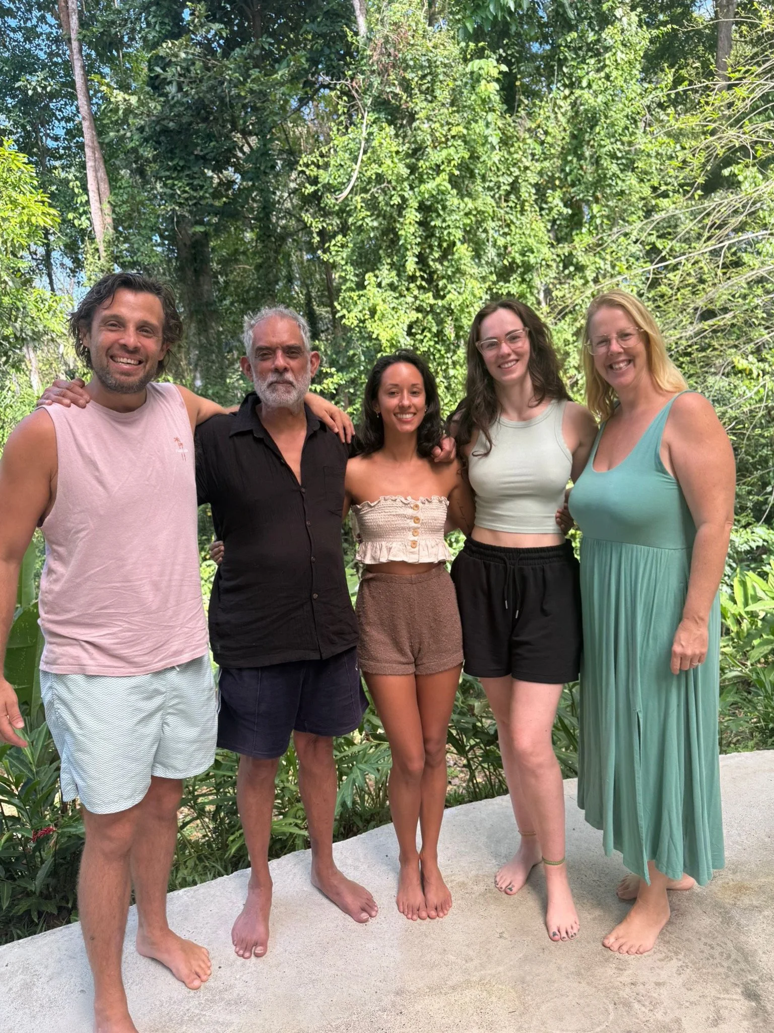 A group of five adults standing in a line, all smiling with green trees in the background