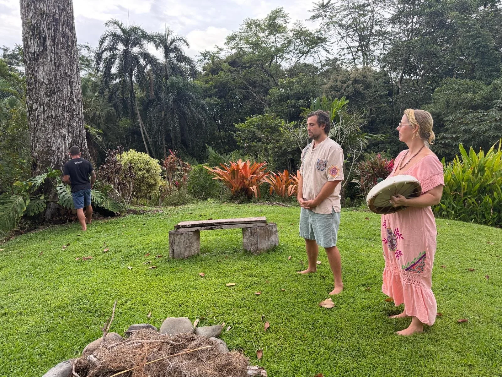 Man and woman stood on grass, wowan holding a drum, jungle background, big tree trunk with man stood in front of it praying