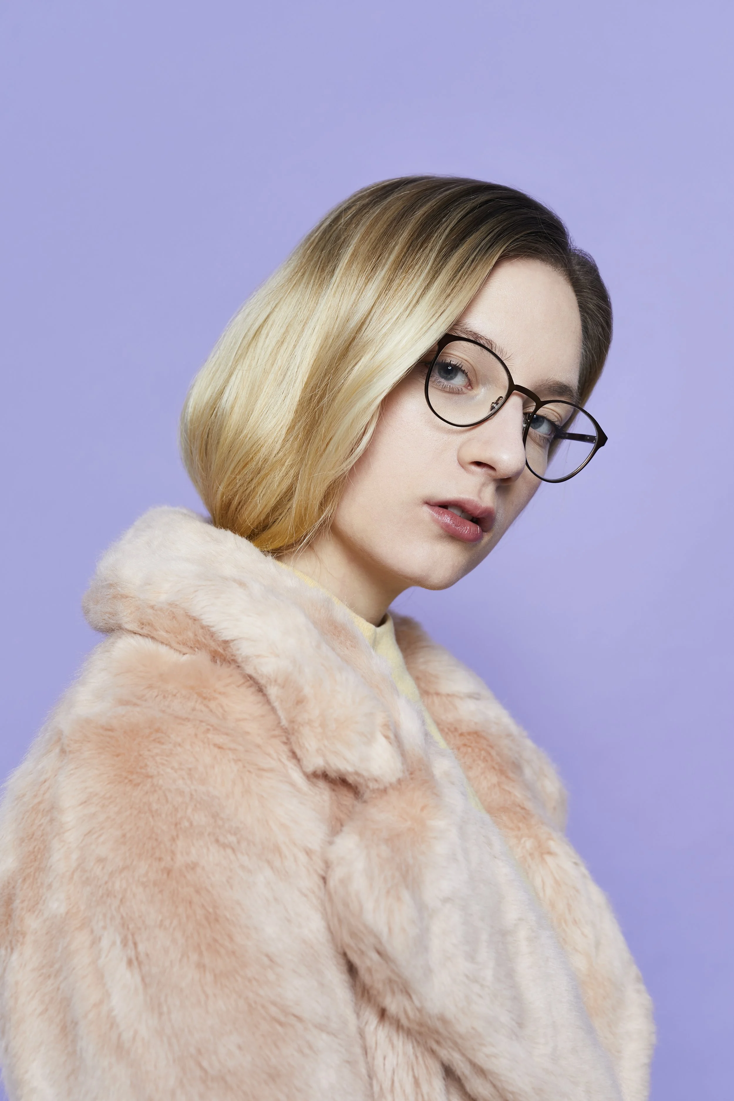 Une femme vêtue d’un manteau de fourrure et portant des lunettes photographiée de côté sur fond violet