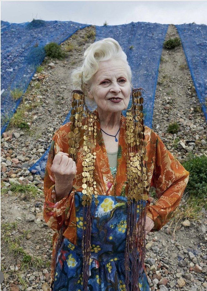 A person with styled white hair wearing a colorful, eclectic outfit featuring a long orange robe with gold circular embellishments and a blue patterned skirt in an outdoor setting.