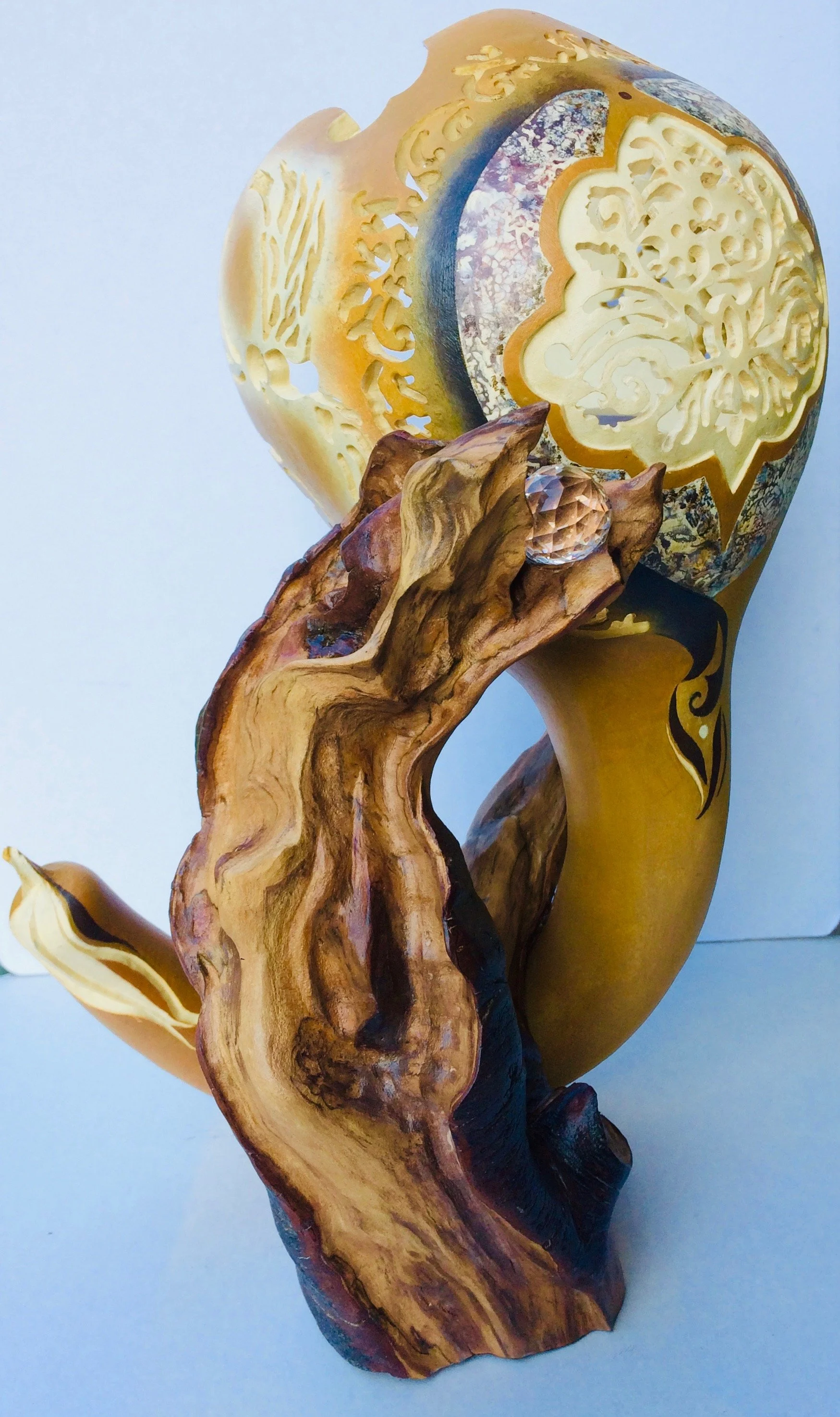 Oeuvre lumineuse raffinée intégrant une calebasse travaillée au Dremel faisant apparaître une rosace sur le devant, fleurs et oiseau sur les côtés.  La coque est peinte et vernie. Le tout repose sur un socle en bois en haut duquel est fixée une boule