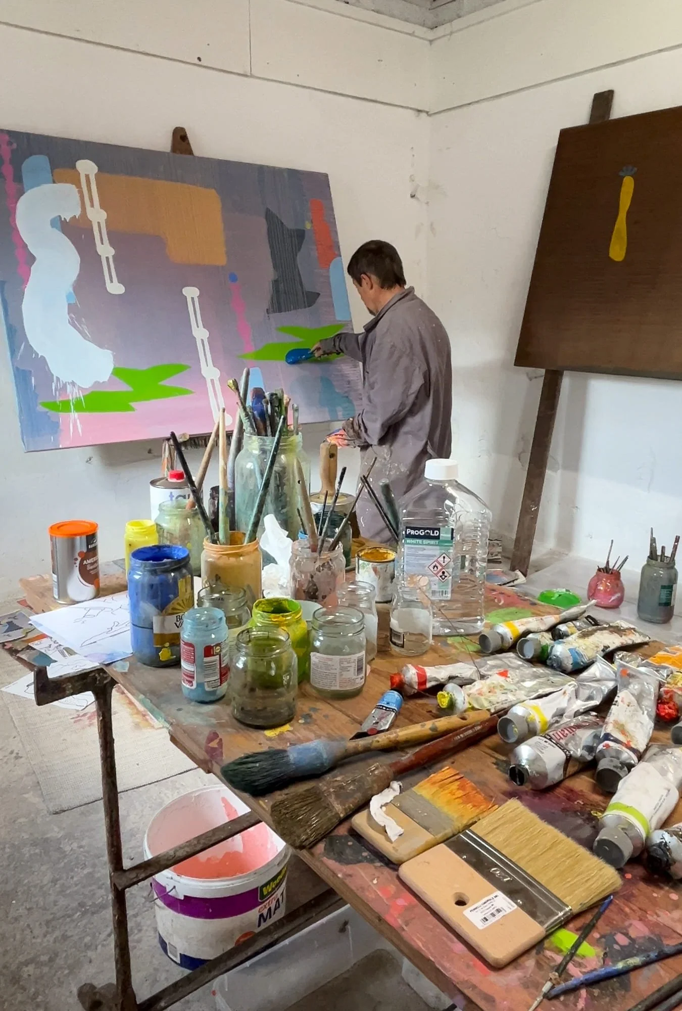 Irish artist painting at canvas in Ballyfin studio, paints brushes and jars covering work table in foreground, large abstract oil in progress with white green pink and grey forms, Martin Finnin