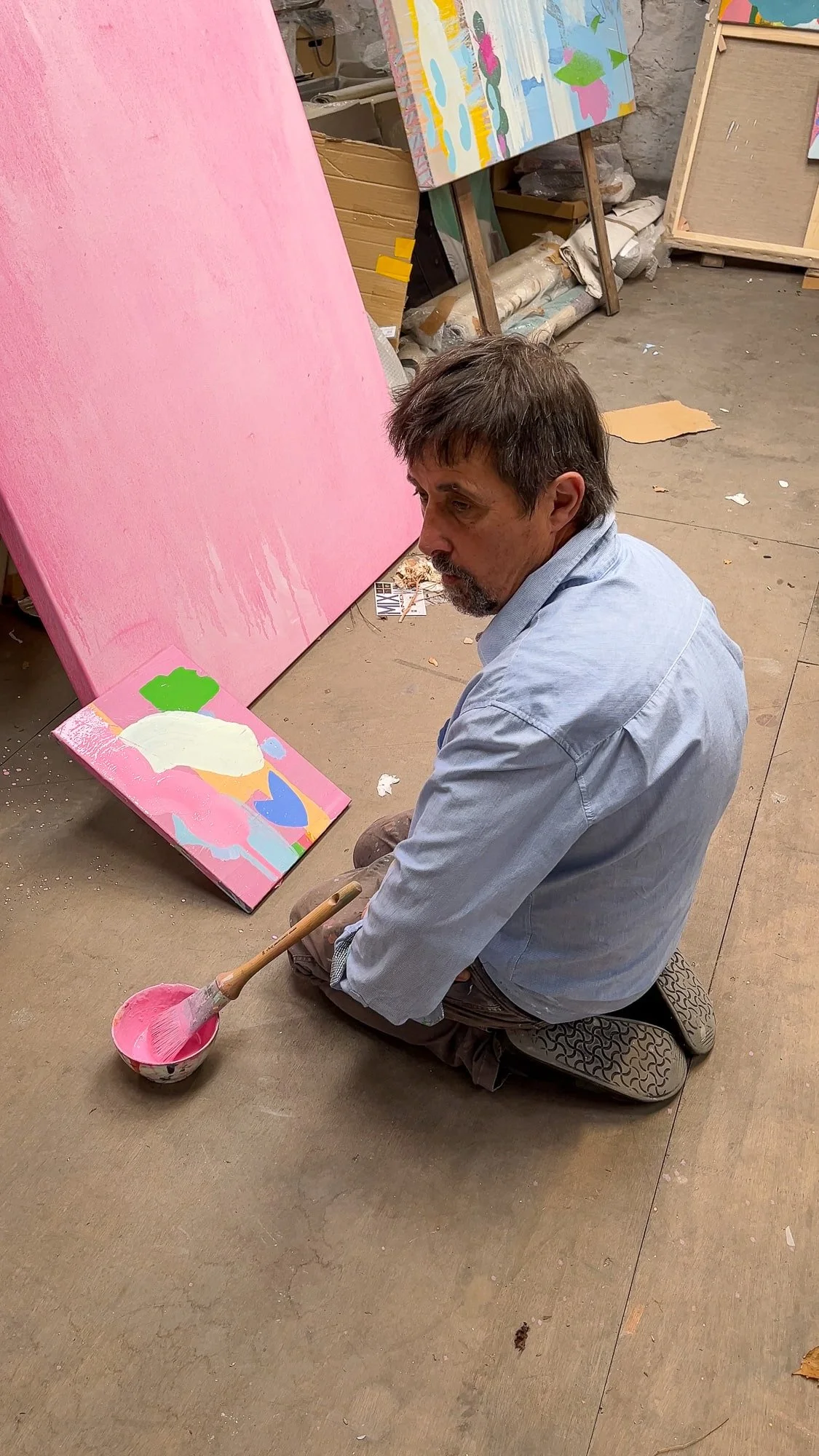 Irish artist seated on studio floor studying small abstract oil painting in progress, pink paint and brush beside him, large pink canvas and other works in background, Martin Finnin West Cork