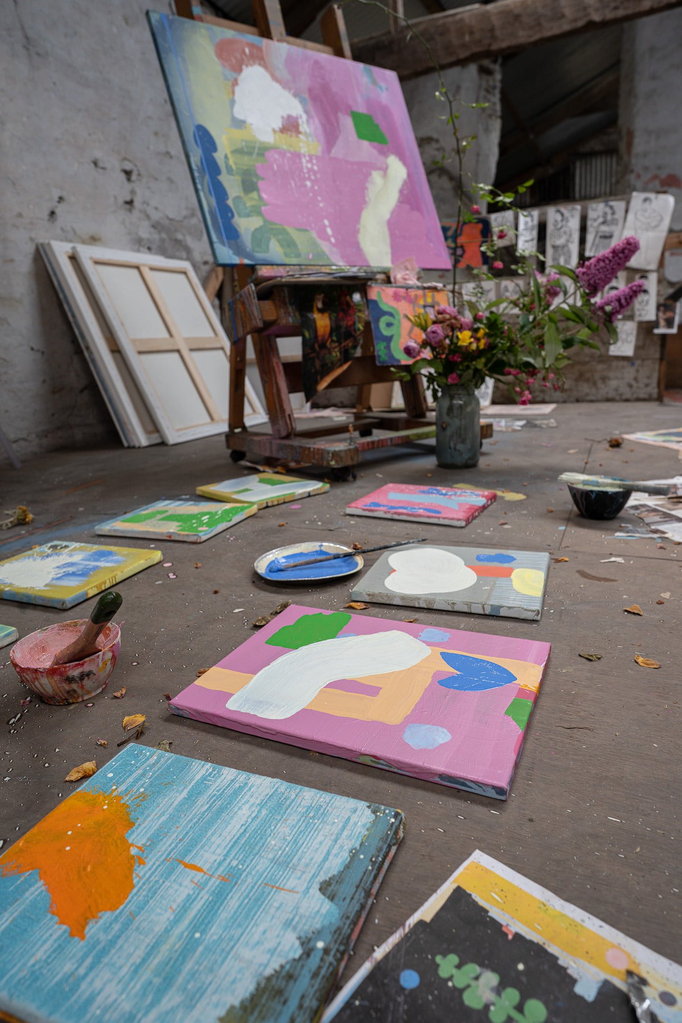 Multiple small abstract oil paintings laid out on West Cork studio floor, pink green blue and orange forms, large canvas on easel and fresh flowers in background, Martin Finnin Irish artist