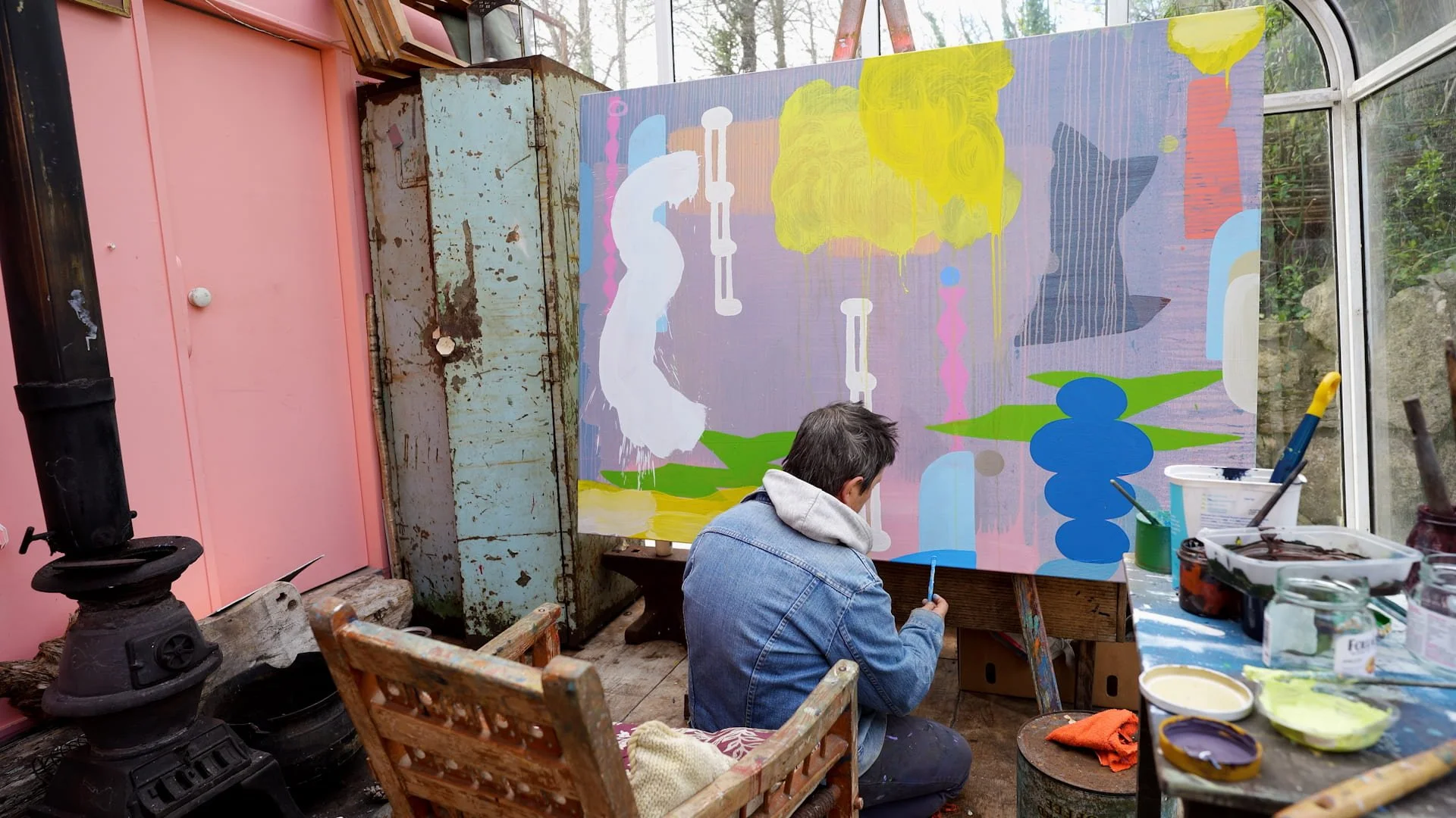 Irish artist kneeling to work on large abstract oil painting in glass studio, yellow green blue and pink forms on lavender ground, paints and jars on table beside him, Martin Finnin West Cork