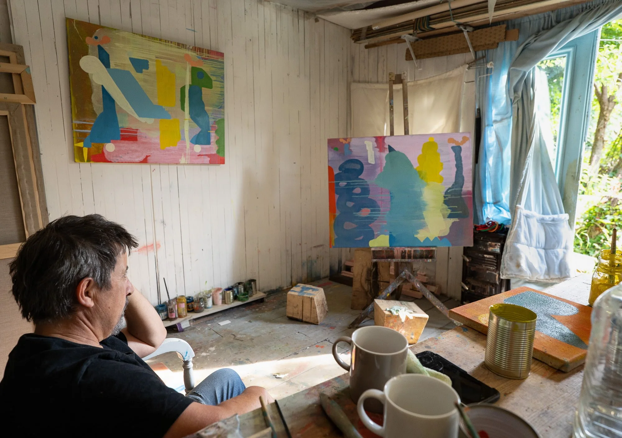 Irish artist in West Cork studio studying two abstract oil paintings, colourful canvases on wall and easel, paint-covered floor and working studio ecology, Martin Finnin