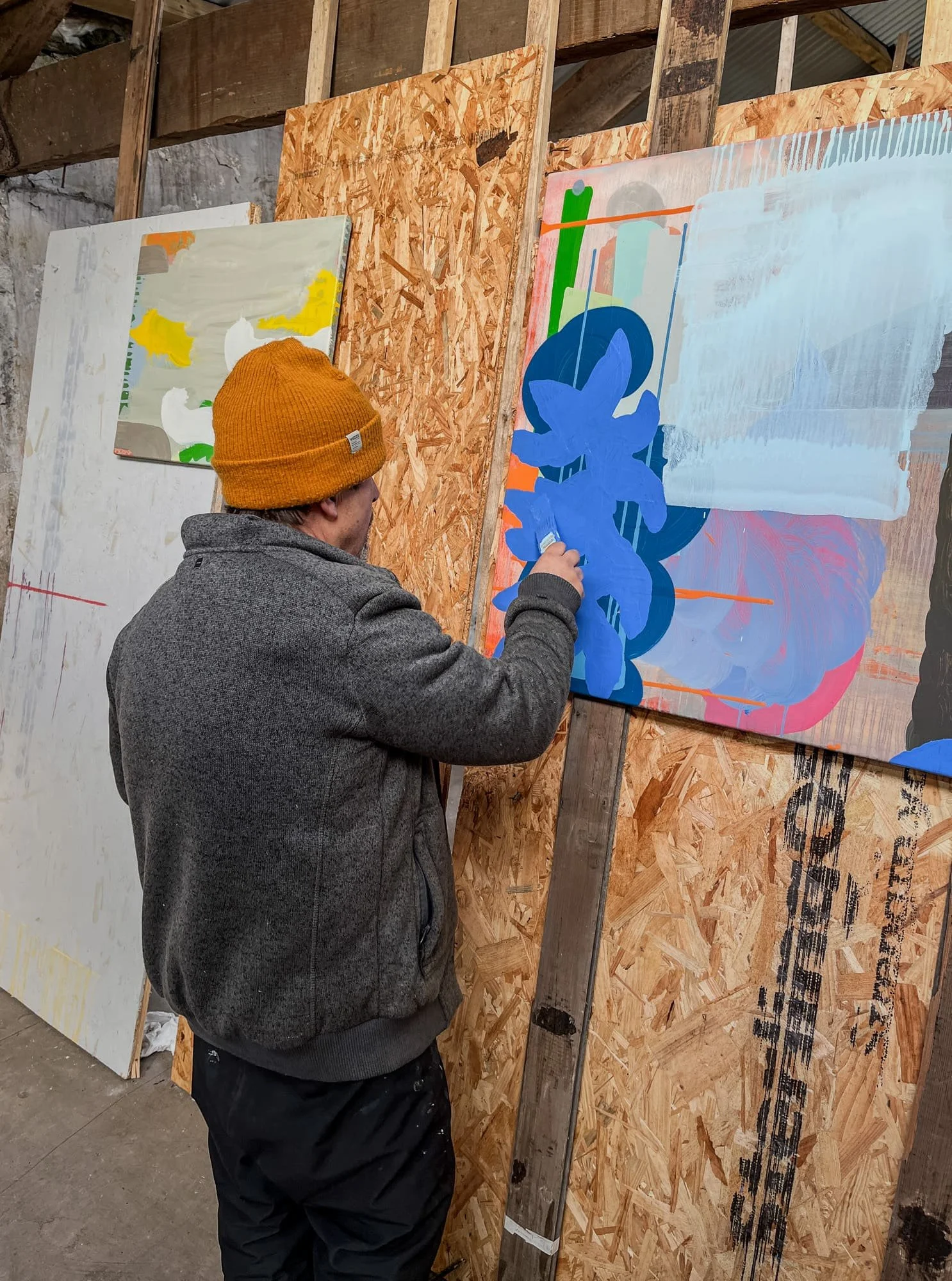 Irish artist Martin Finnin painting bold blue forms onto abstract canvas leaning against studio wall, multiple canvases visible in background, oil painting in progress in West Cork studio