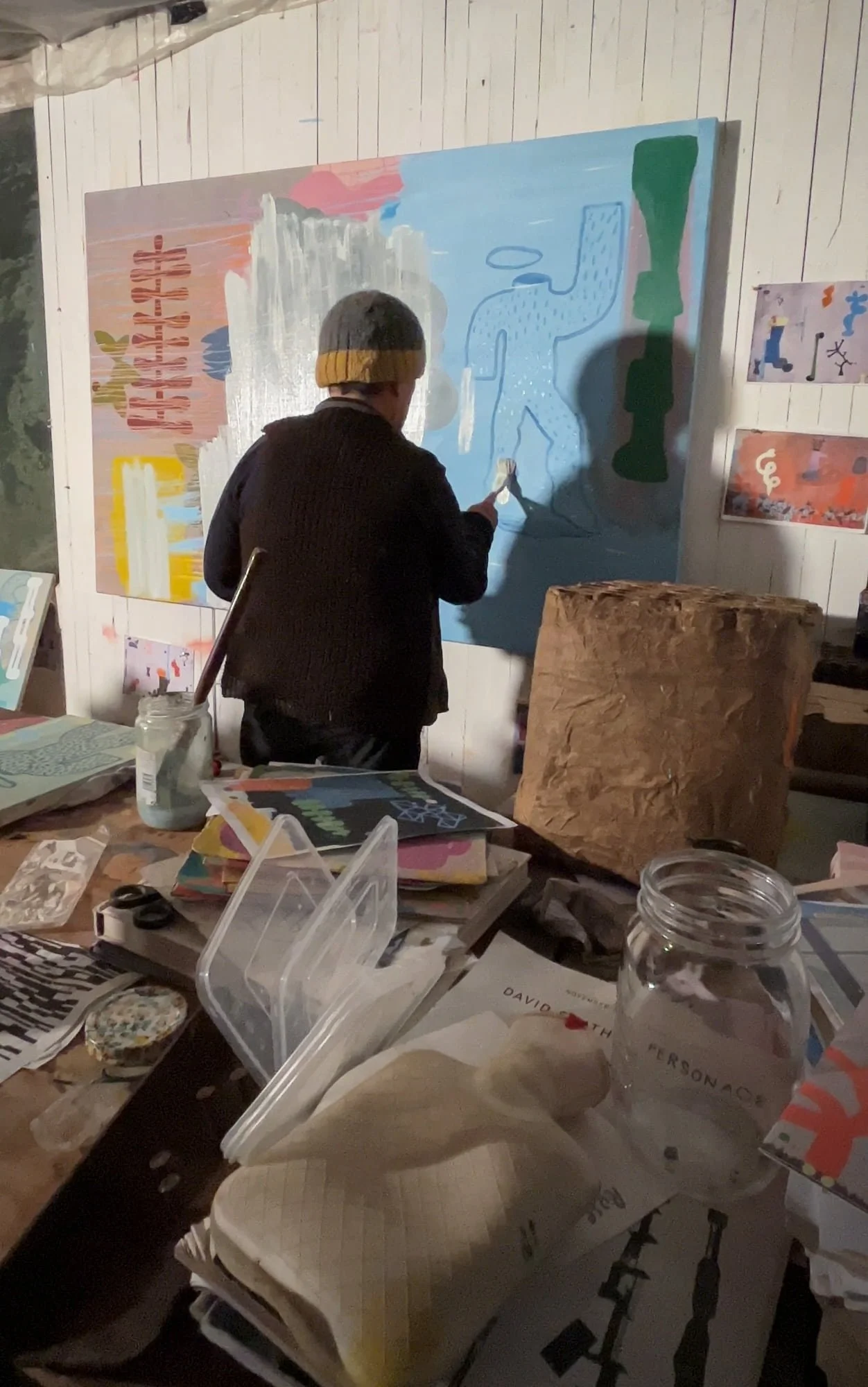 Irish artist painting outlined figure onto blue ground of large canvas, pink and green forms on left, source material and books covering studio table in foreground, Martin Finnin West Cork