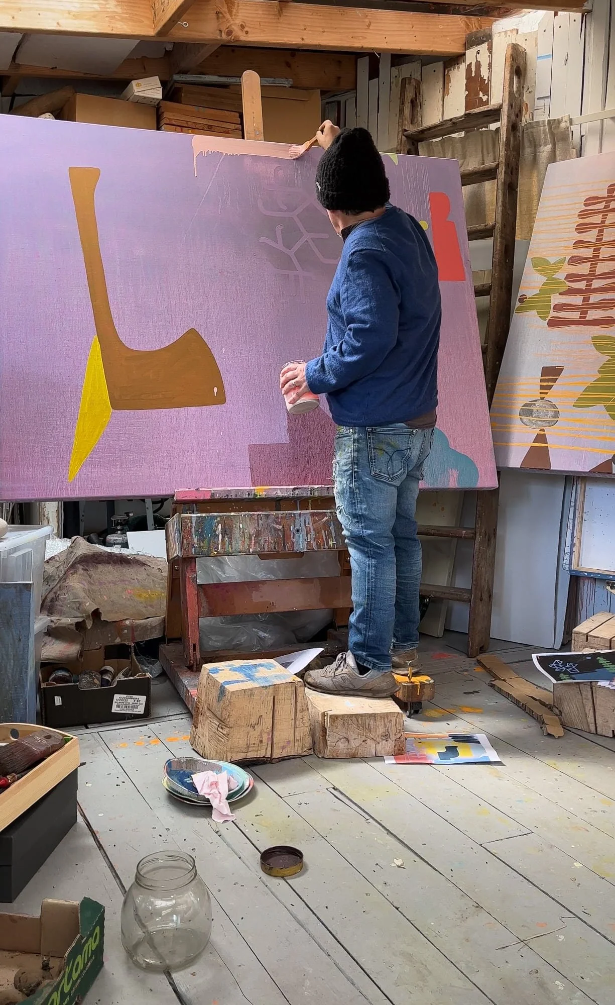 Irish artist painting upper section of large canvas on easel, lavender pink ground with ochre yellow and terracotta forms developing, West Cork studio with source material on floor, Martin Finnin