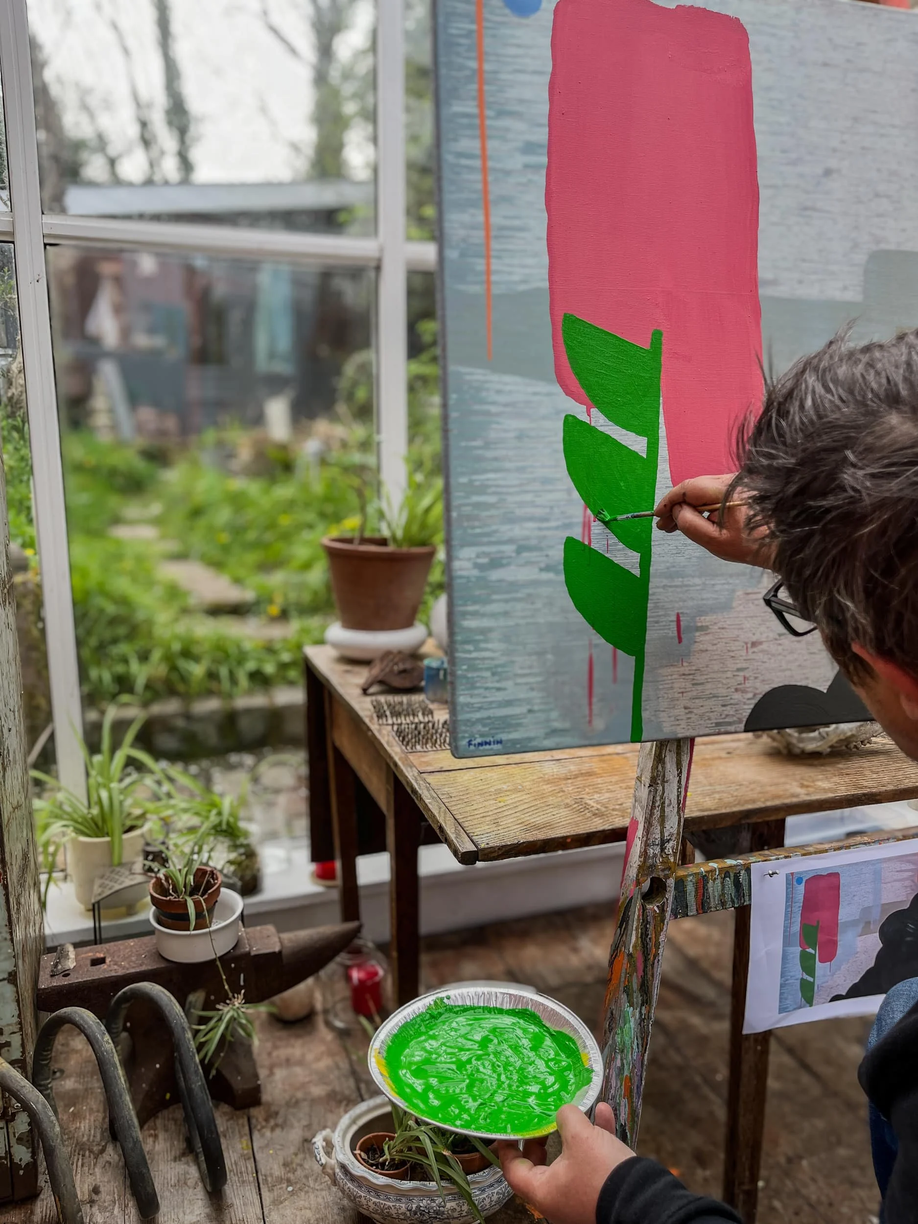 Irish artist Martin Finnin painting bright green stem onto abstract canvas in greenhouse studio with garden view, holding palette of green paint, plants and garden visible through glass, West Cork
