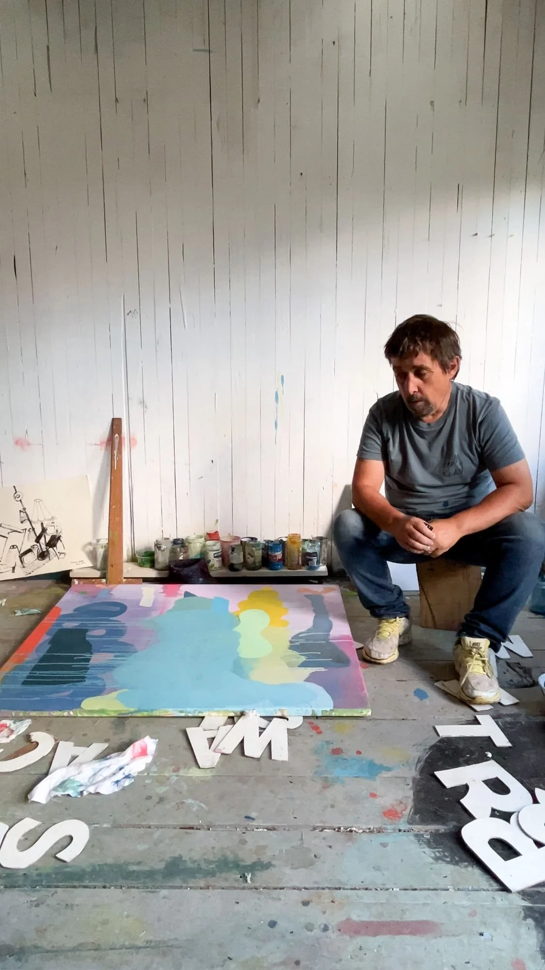 Irish artist Martin Finnin sitting beside abstract canvas on studio floor, blue teal and yellow forms developing, large cut-out letters scattered around, painting in progress, artist studio