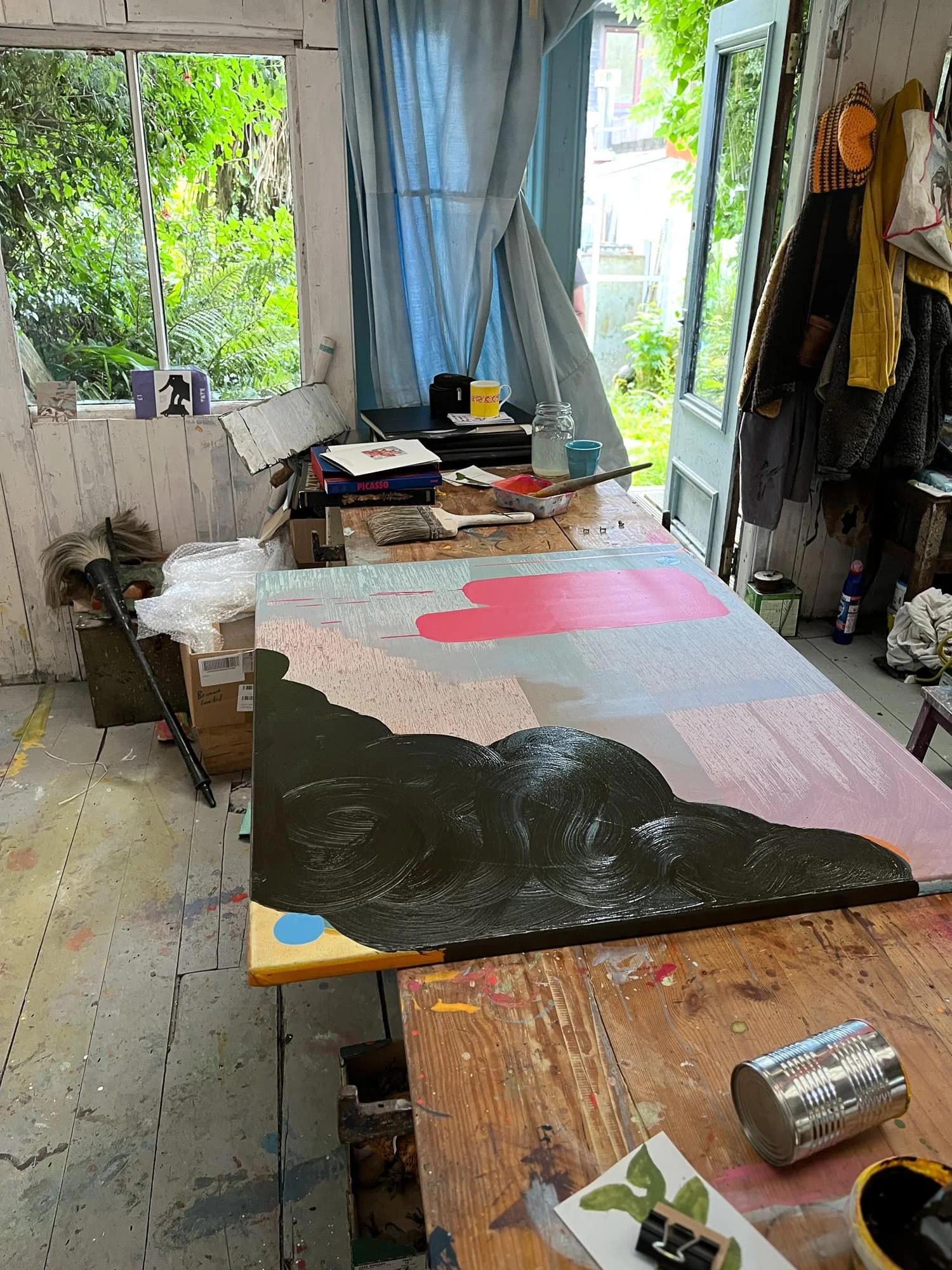 Abstract oil painting on studio table with dark black cloud form and pink shape developing, lush garden visible through open studio door, books brushes and paint tins on worktable, Martin Finnin West Cork studio