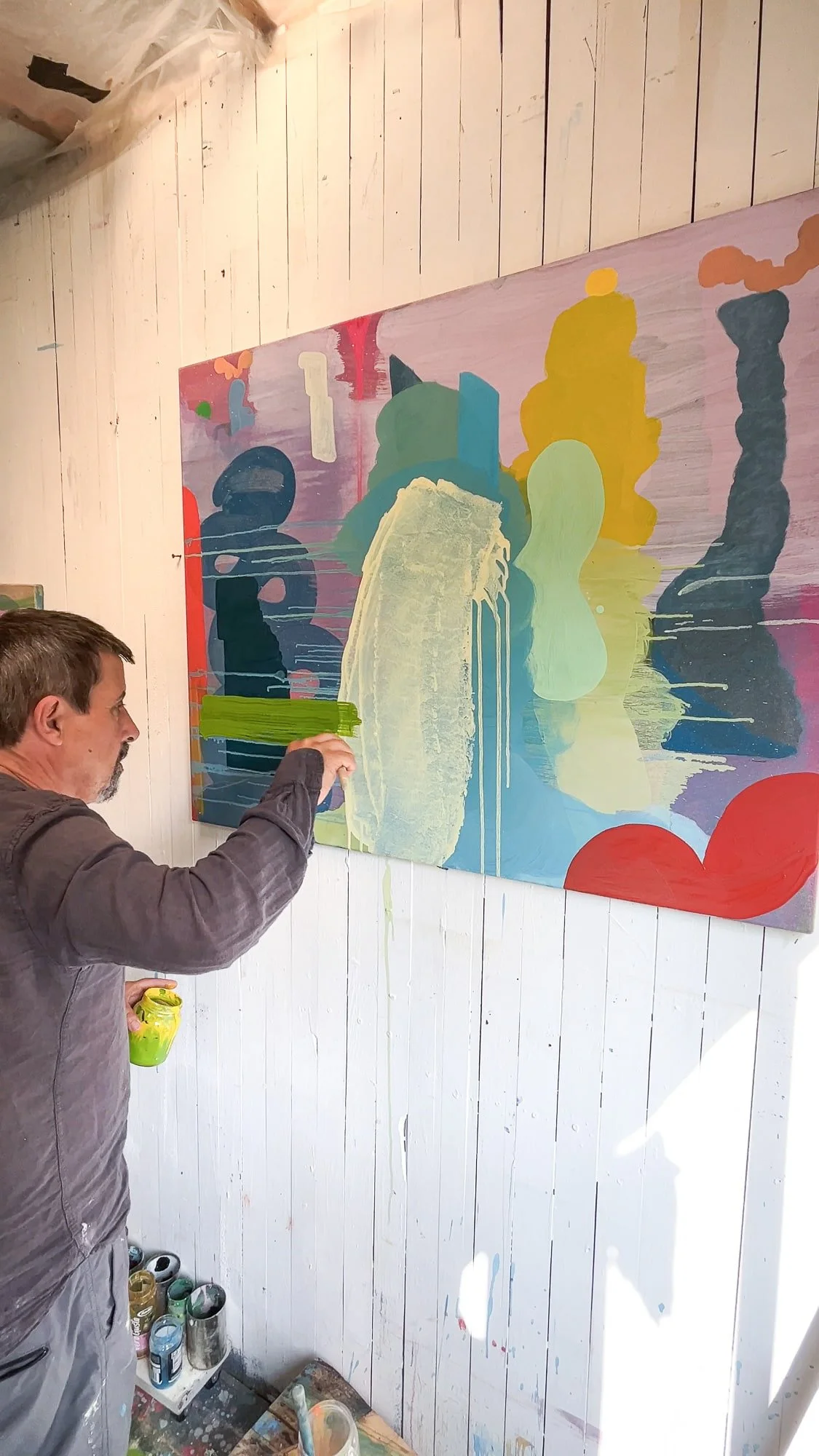 Irish artist Martin Finnin applying pale green paint with brush to large abstract canvas on studio wall, vivid multicolour oil painting with yellow blue and red forms in progress, West Cork studio