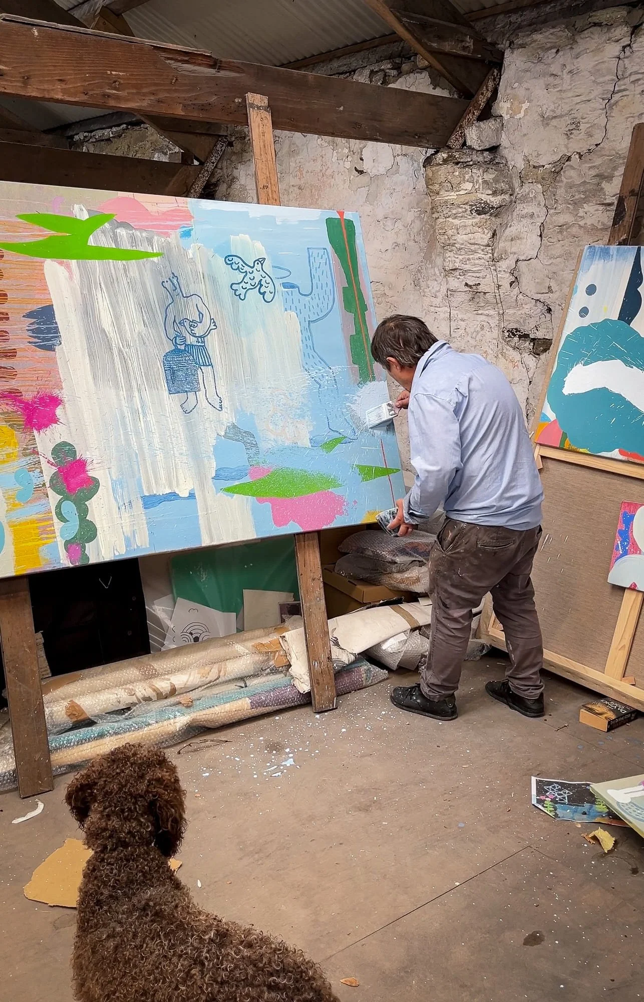 Irish artist working on large abstract canvas on easel in West Cork studio, blue green pink and white forms with drawn figures, dog watching from foreground, Martin Finnin Irish artist