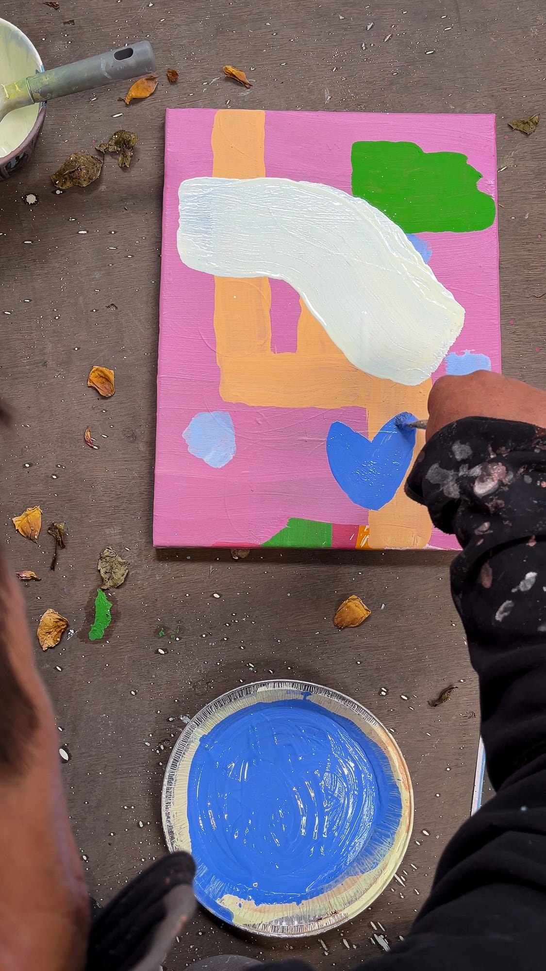 Artist's hand adding blue form to small abstract oil painting on studio floor, white green orange and pink forms developing, blue paint in dish beside canvas, Martin Finnin West Cork