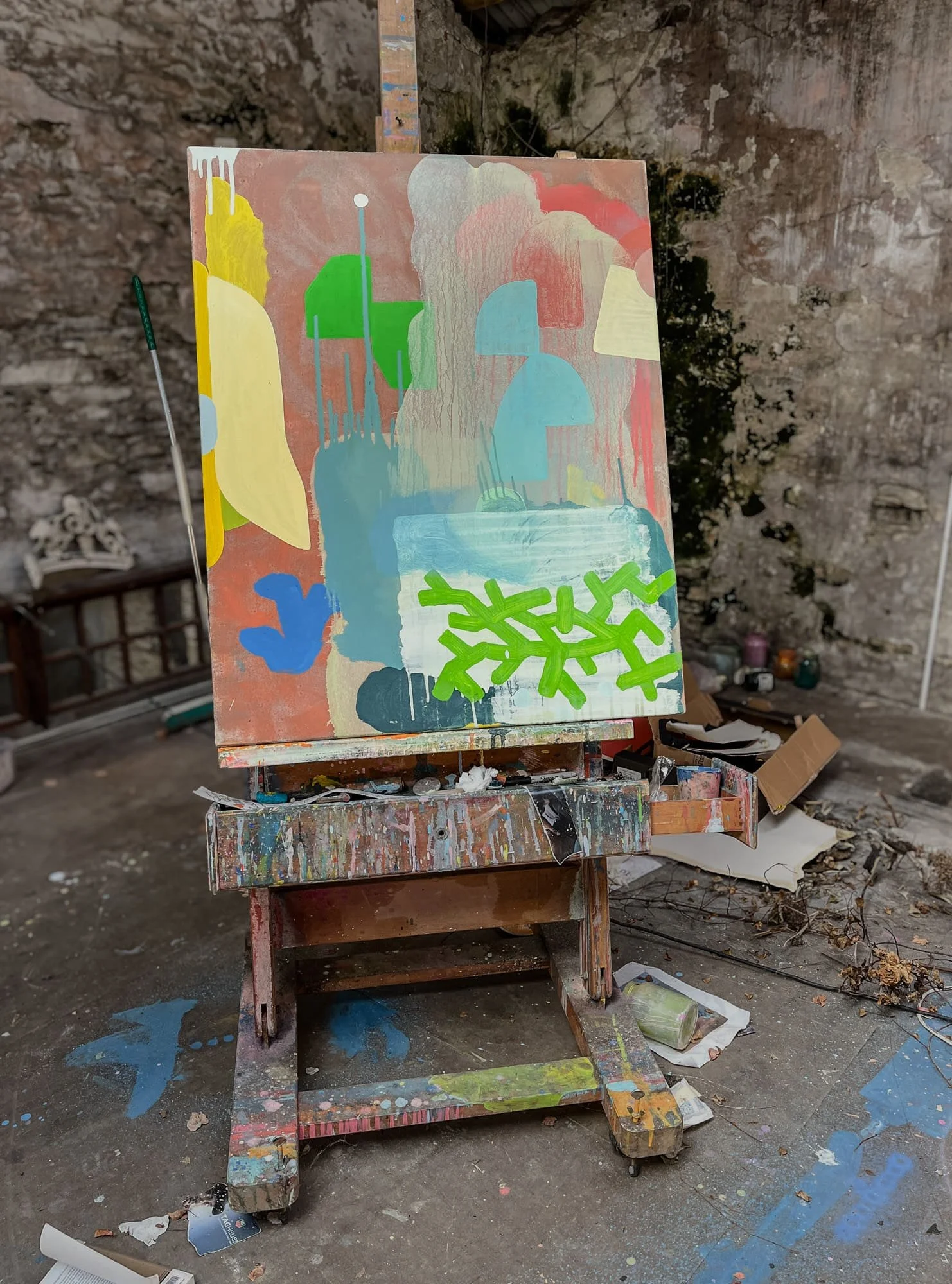 Abstract oil painting in progress on paint-covered easel in stone-walled West Cork studio, yellow green blue and terracotta forms with bright green marks emerging, Martin Finnin Irish artist