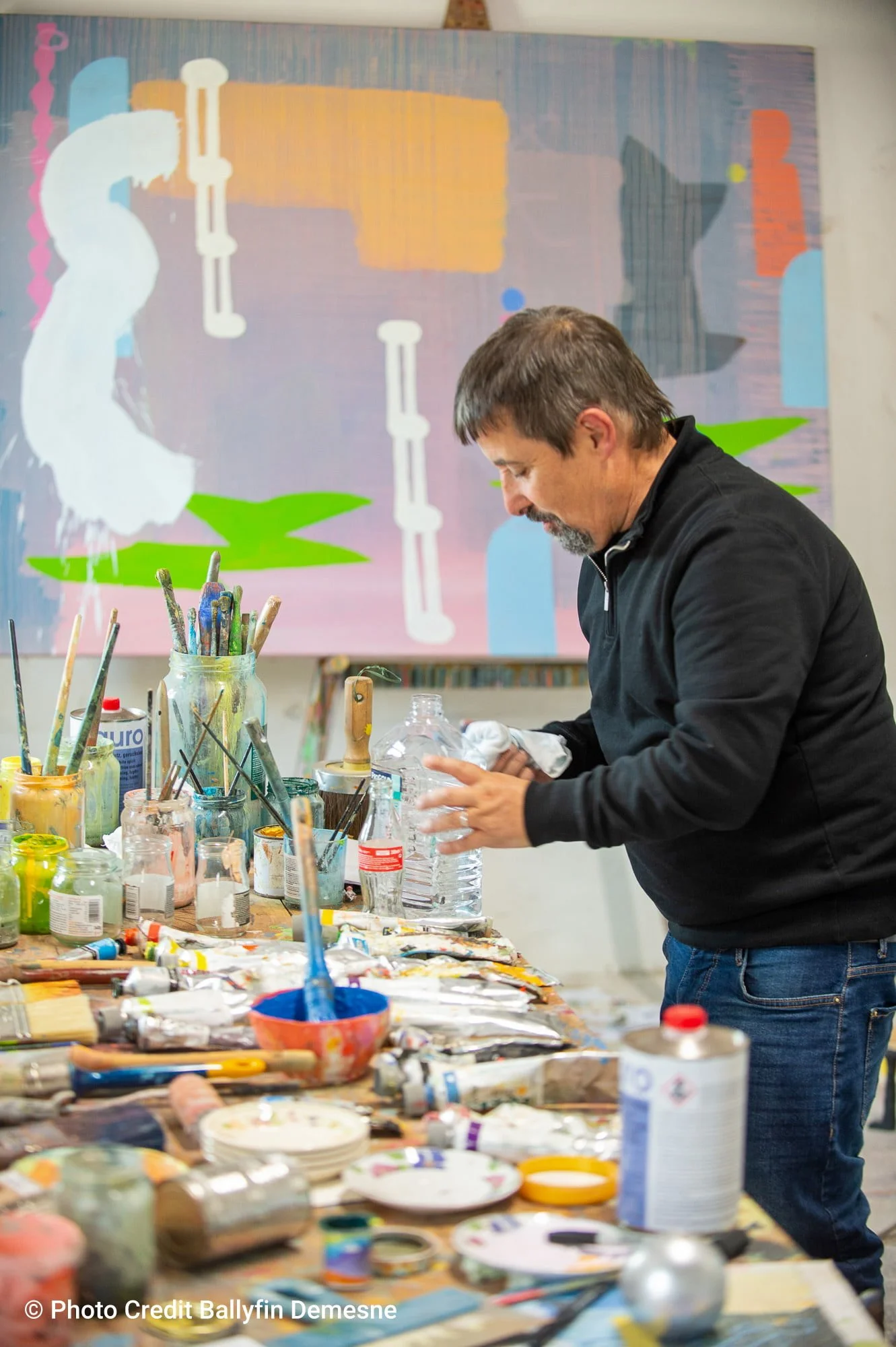 Irish artist at work table in Ballyfin studio, large abstract oil painting in progress behind him, paints brushes and materials covering the table, Martin Finnin