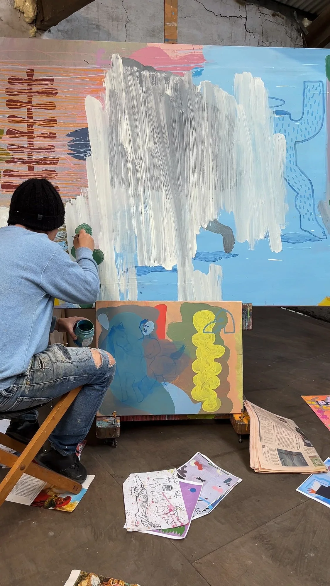 Irish artist seated painting white waterfall passage onto large canvas, blue and pink ground developing, smaller figurative painting on easel below, source material scattered on studio floor, Martin Finnin West Cork