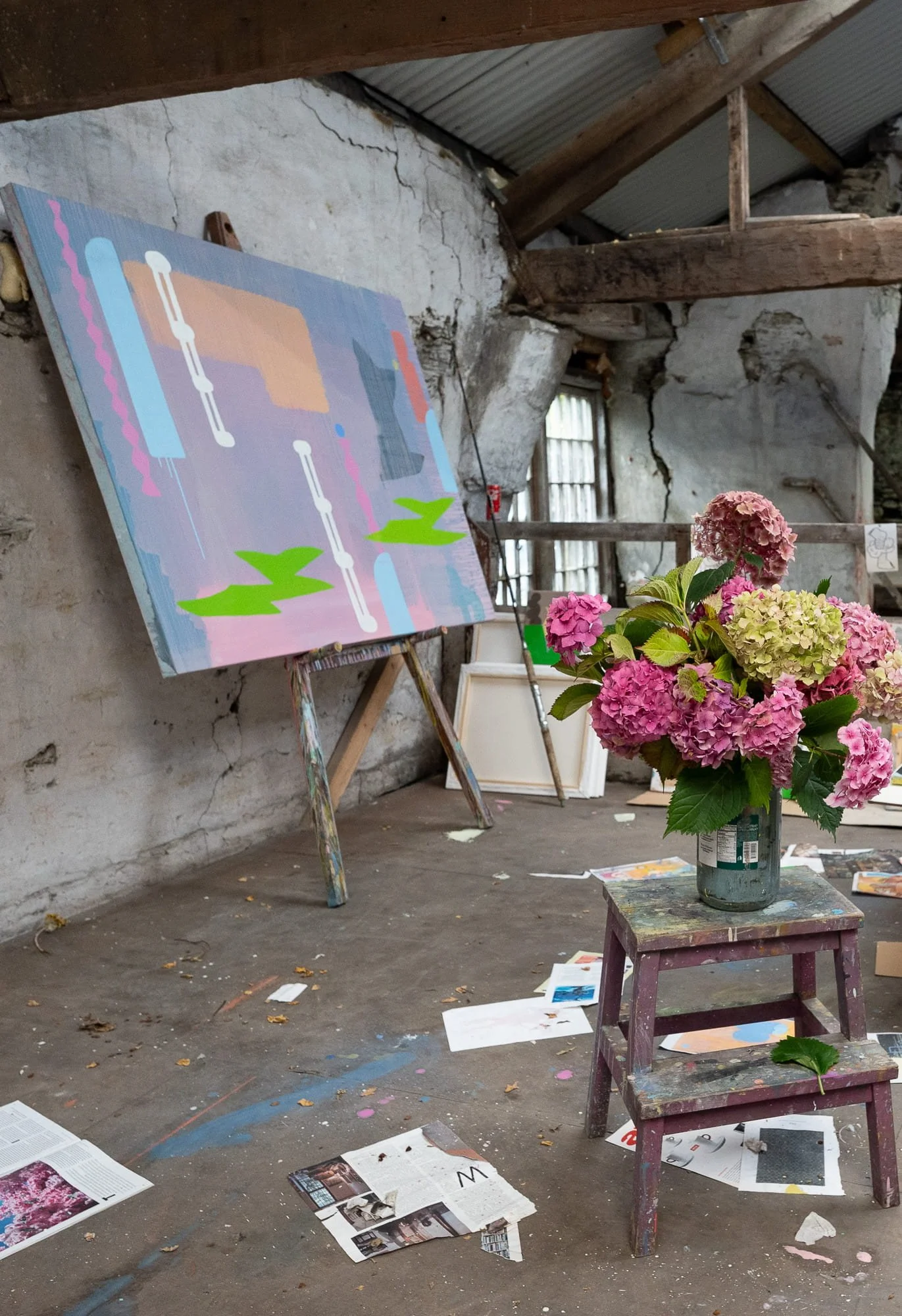 Abstract oil painting on easel in West Cork studio, lavender pink blue and green forms in progress, fresh pink hydrangeas in foreground and source material on floor, Martin Finnin Irish artist