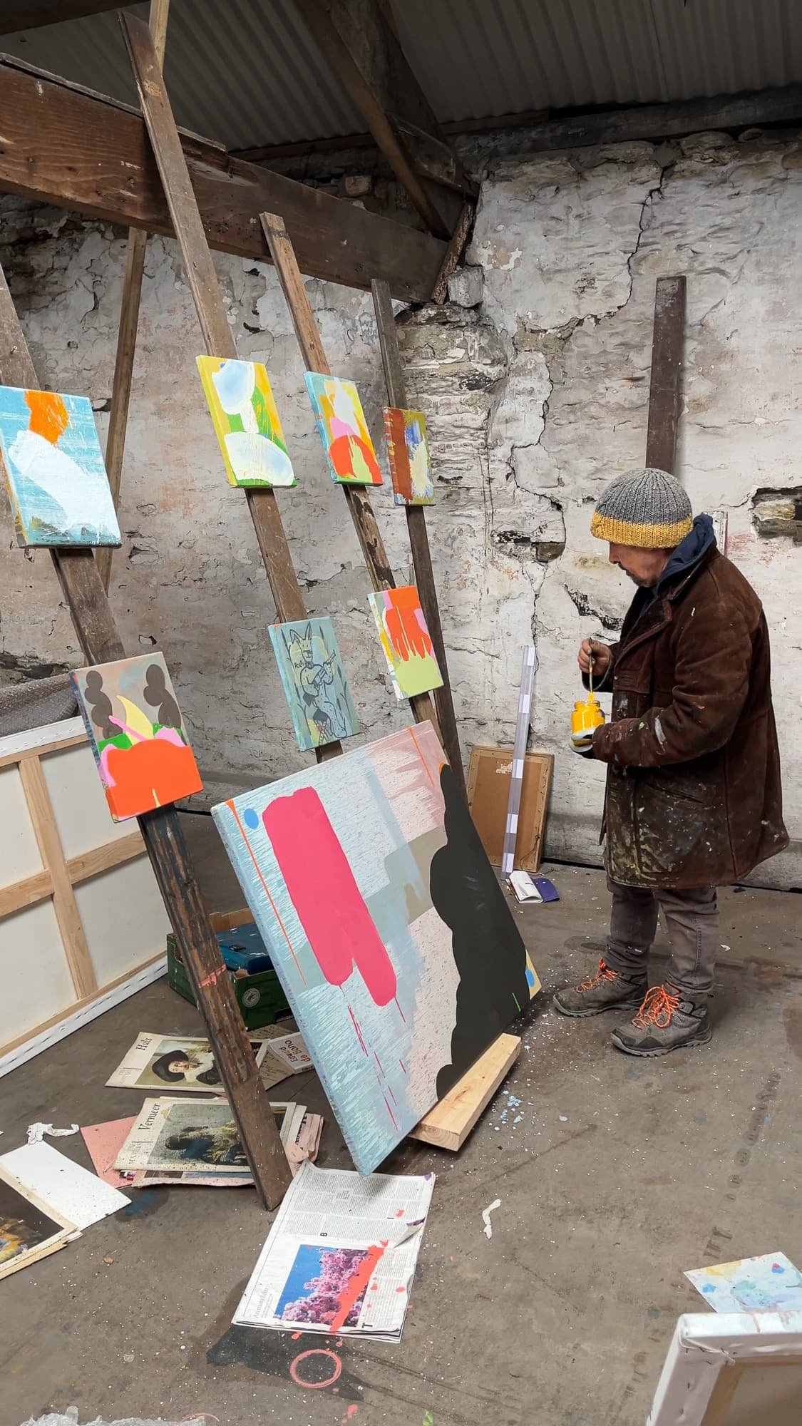 Irish artist Martin Finnin standing in studio surrounded by multiple small canvases on easel rack, large abstract painting with pink and black forms on studio floor, newspapers and art books scattered around