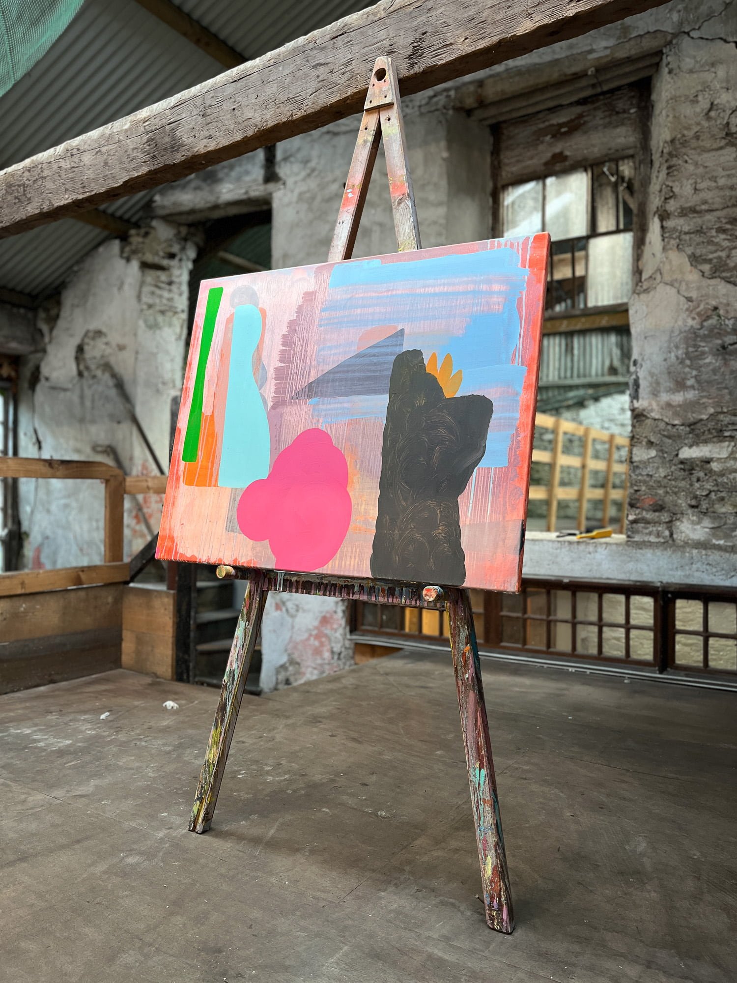 Abstract oil painting in progress on easel in raw stone West Cork studio, coral pink canvas with bold pink blue and dark black forms developing, paint-covered easel in large studio space, Martin Finnin