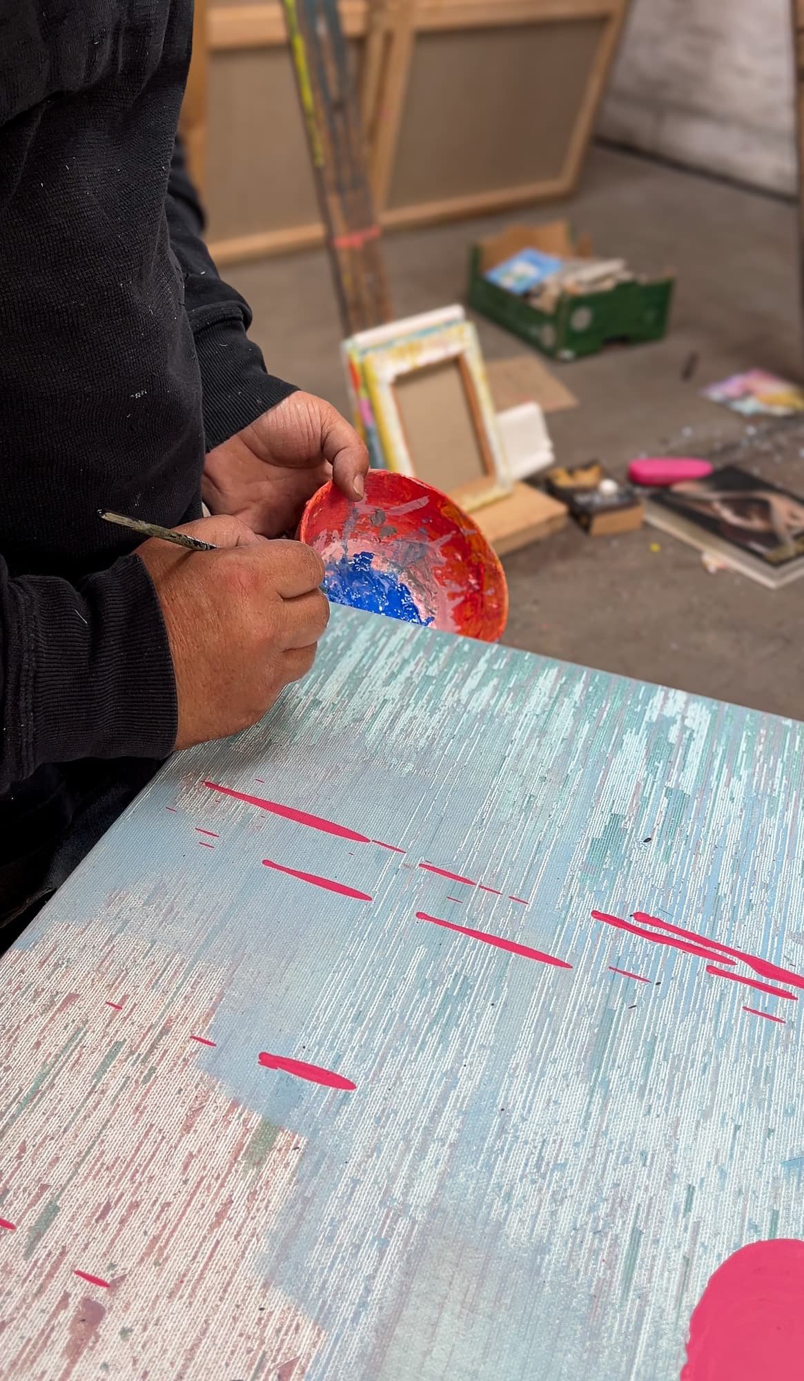Irish artist mixing paint over canvas in progress, pale blue ground with raw silk texture and pink splash marks emerging, West Cork studio, Martin Finnin