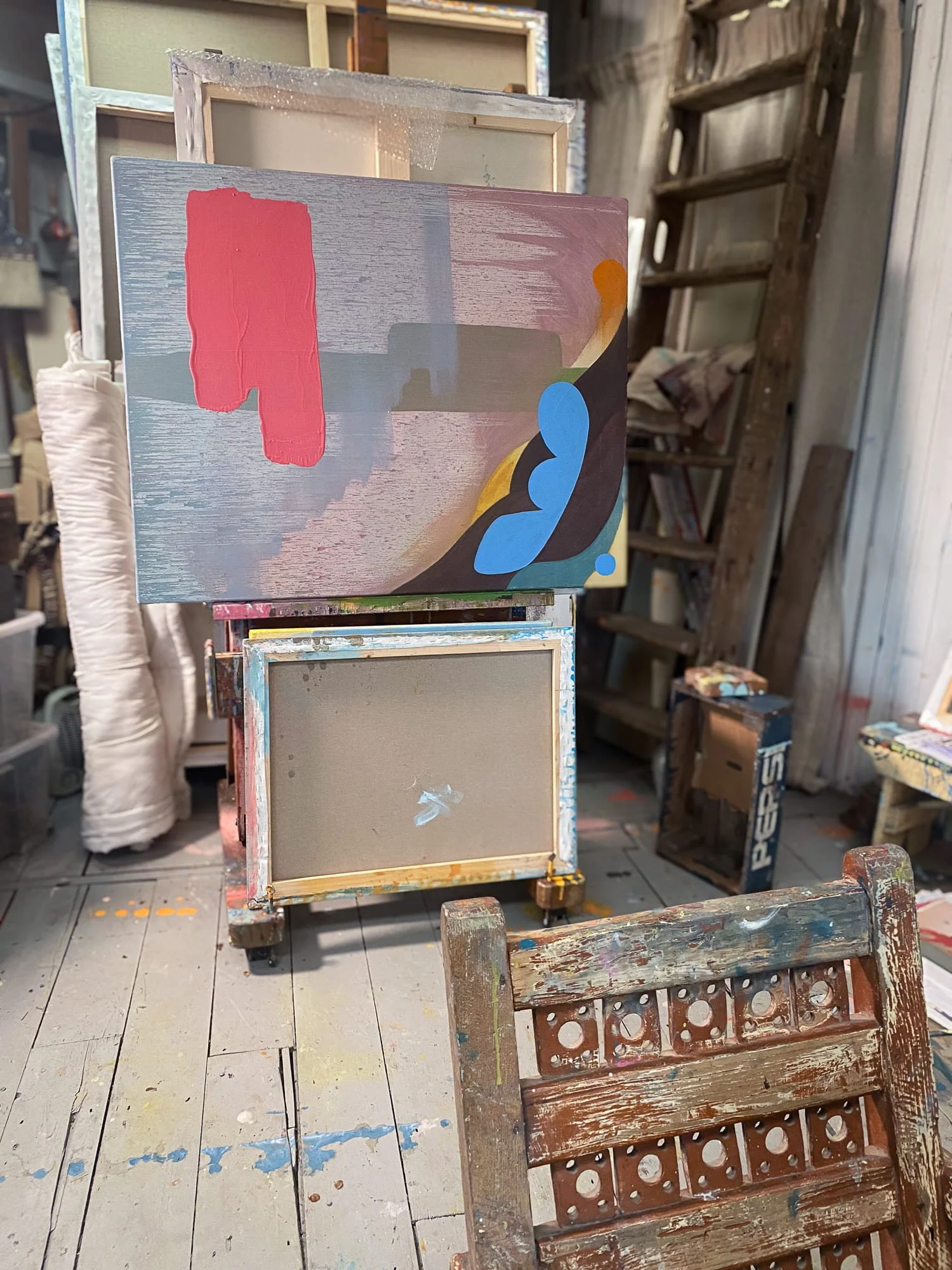 Early stage abstract oil painting on studio easel, red and mauve forms on pale grey canvas with blue and dark brown emerging, accumulated studio objects and wooden ladder visible, Martin Finnin West Cork