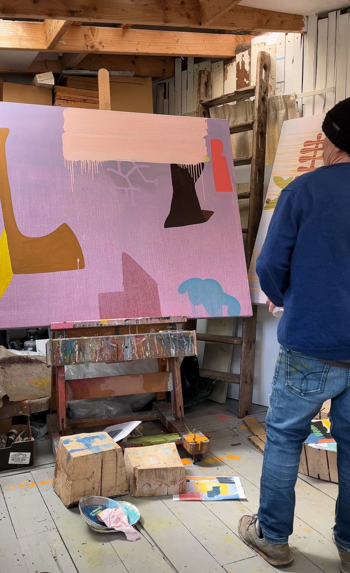 Early stage abstract oil painting on easel in West Cork studio, lavender pink ground with dark brown ochre and blue forms emerging, artist standing beside canvas, Martin Finnin Irish artist