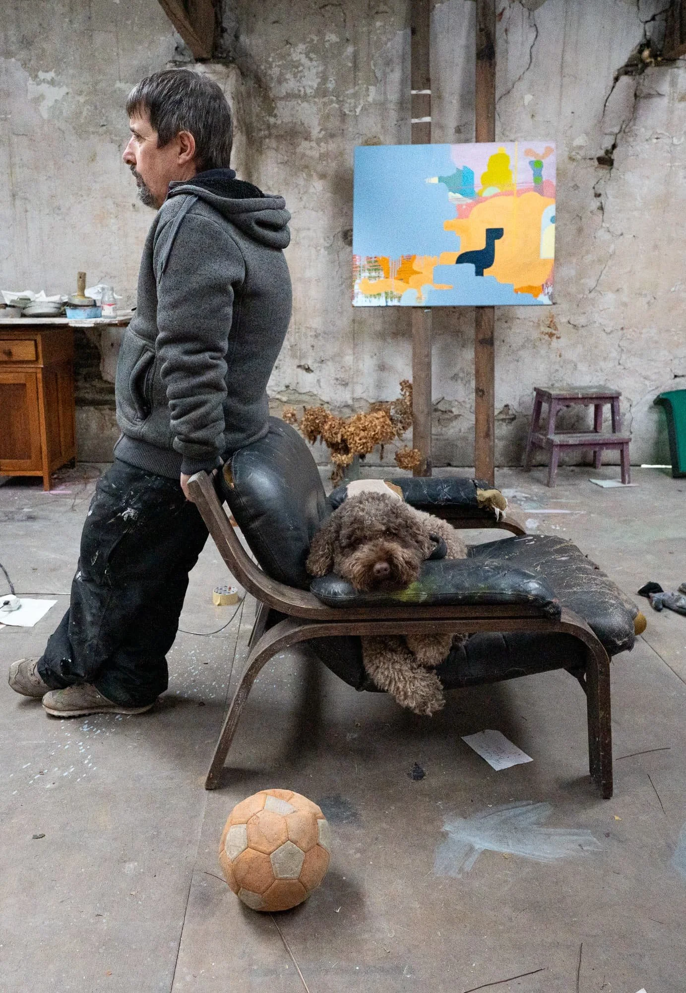 Irish artist Martin Finnin standing in stone West Cork studio with dog resting on chair and football on floor, abstract oil painting visible on easel in background, studio life