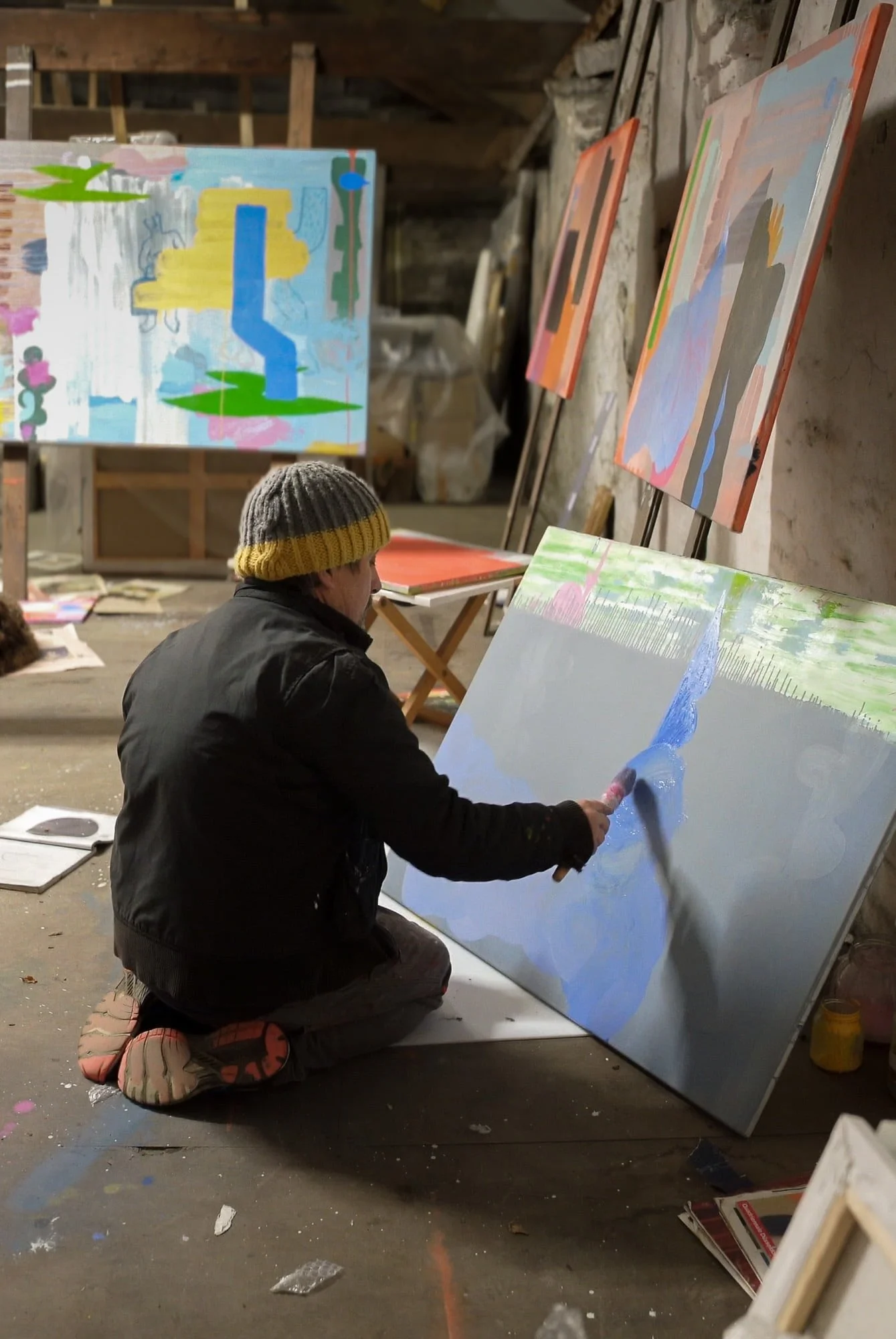 Irish artist Martin Finnin kneeling on studio floor painting blue form onto canvas, multiple abstract paintings visible in background of studio, painting in progress