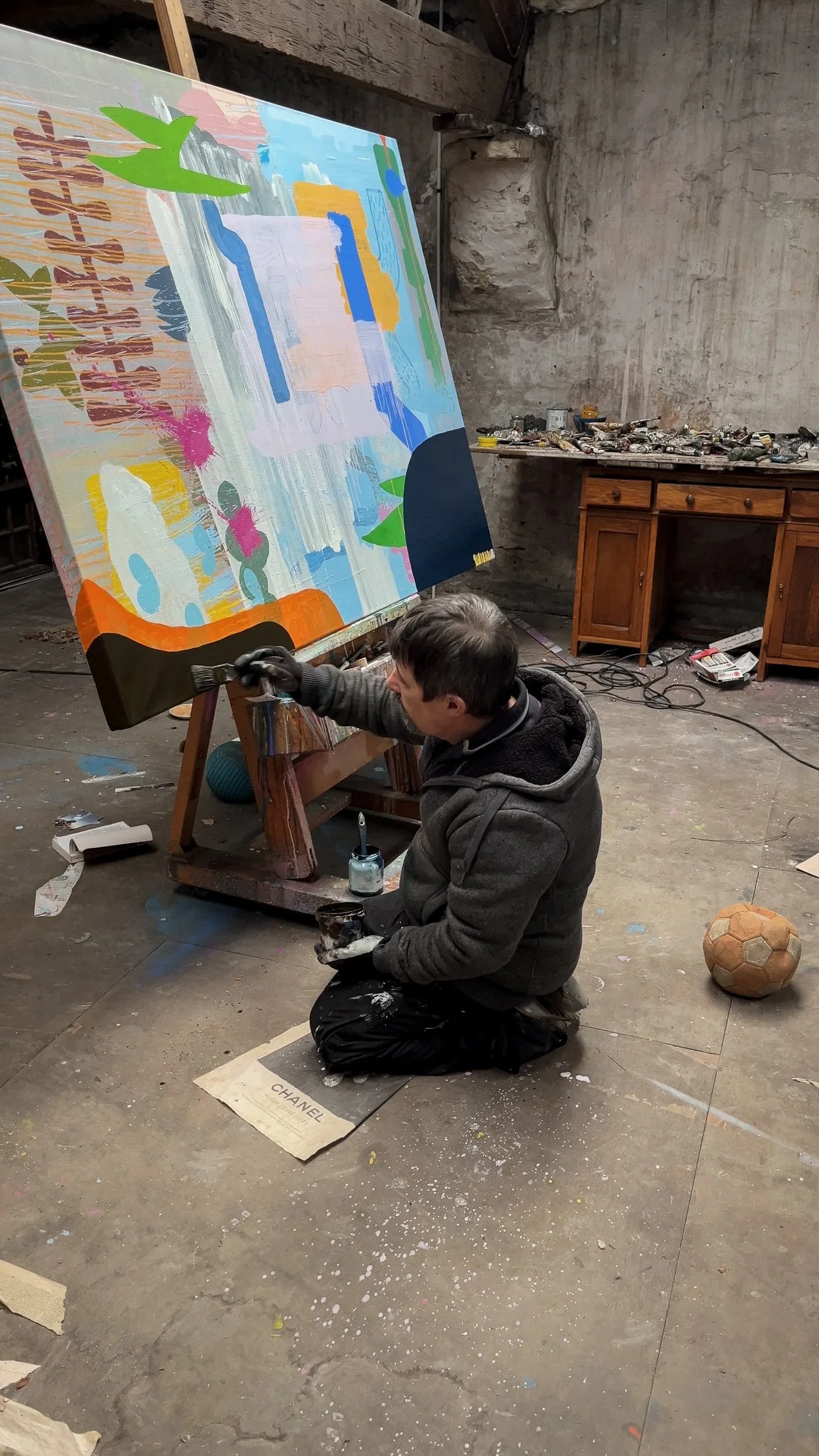 Irish artist kneeling to paint lower section of large abstract canvas on easel, blue green orange and navy forms, West Cork stone-walled studio with old football and antique desk, Martin Finnin