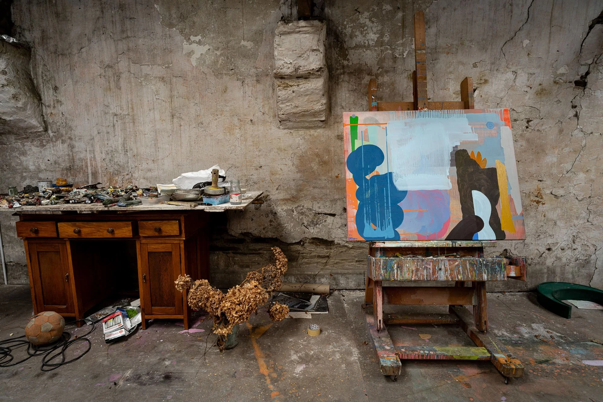Abstract oil painting on easel in West Cork studio with paint-covered workbench, dried hydrangeas, old football and accumulated studio objects, chance chamber of Irish artist Martin Finnin