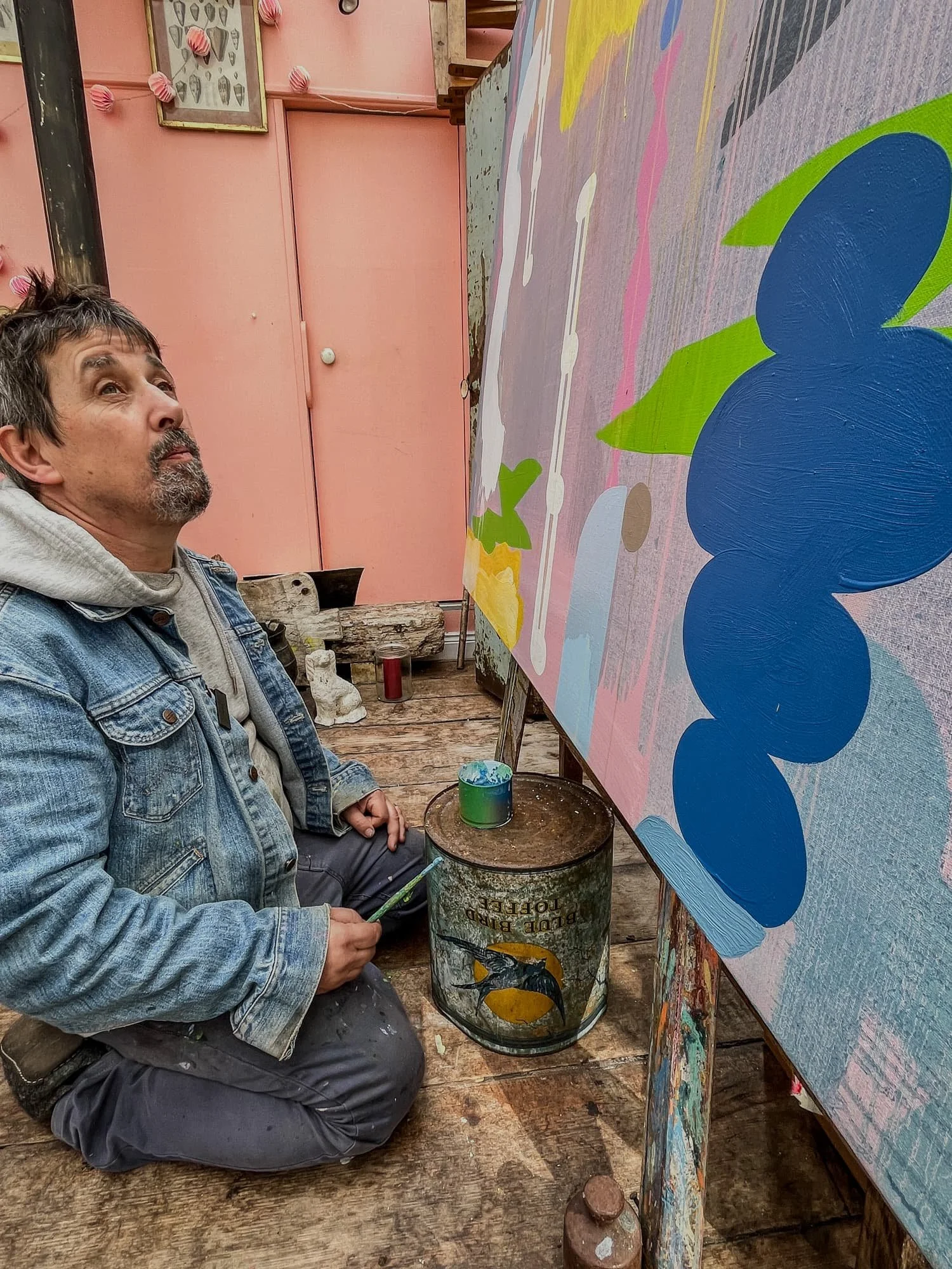 Irish artist seated on studio floor looking up at large abstract oil painting, blue green and pink forms on lavender ground, West Cork studio with found objects, Martin Finnin