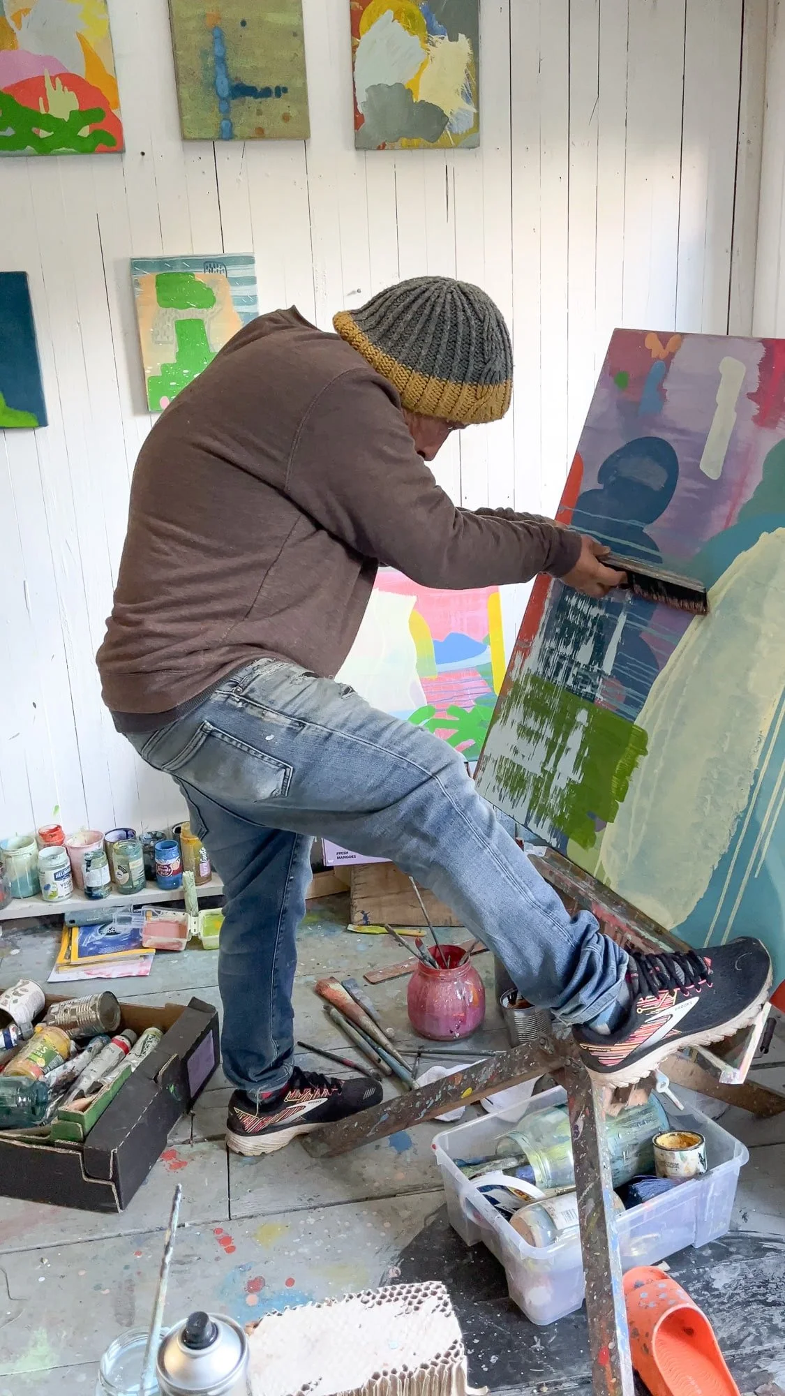 Irish artist Martin Finnin working intensely on abstract canvas balanced on studio table, multiple paintings visible on wall behind, paint-covered studio floor with brushes and pigments, West Cork