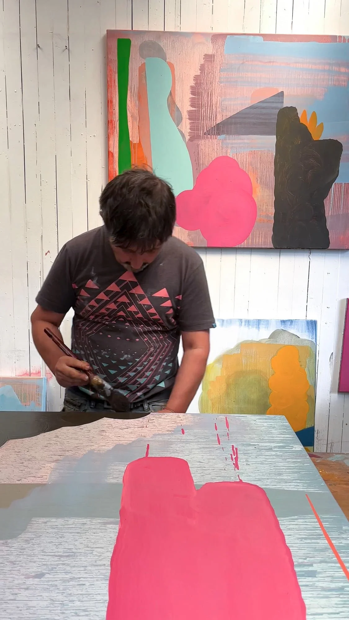 Irish artist Martin Finnin working on abstract canvas on studio table, bold pink forms emerging, multiple paintings in progress visible on white panelled wall behind, West Cork studio