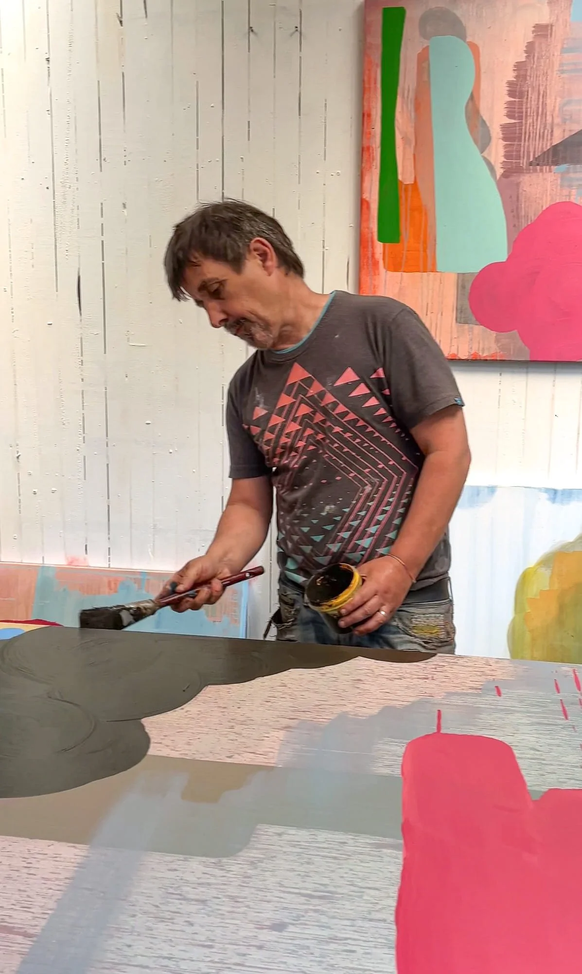 Irish artist Martin Finnin painting dark cloud form onto large abstract canvas on studio floor, pink and grey forms visible, multiple paintings in progress on white panelled studio wall behind, West Cork