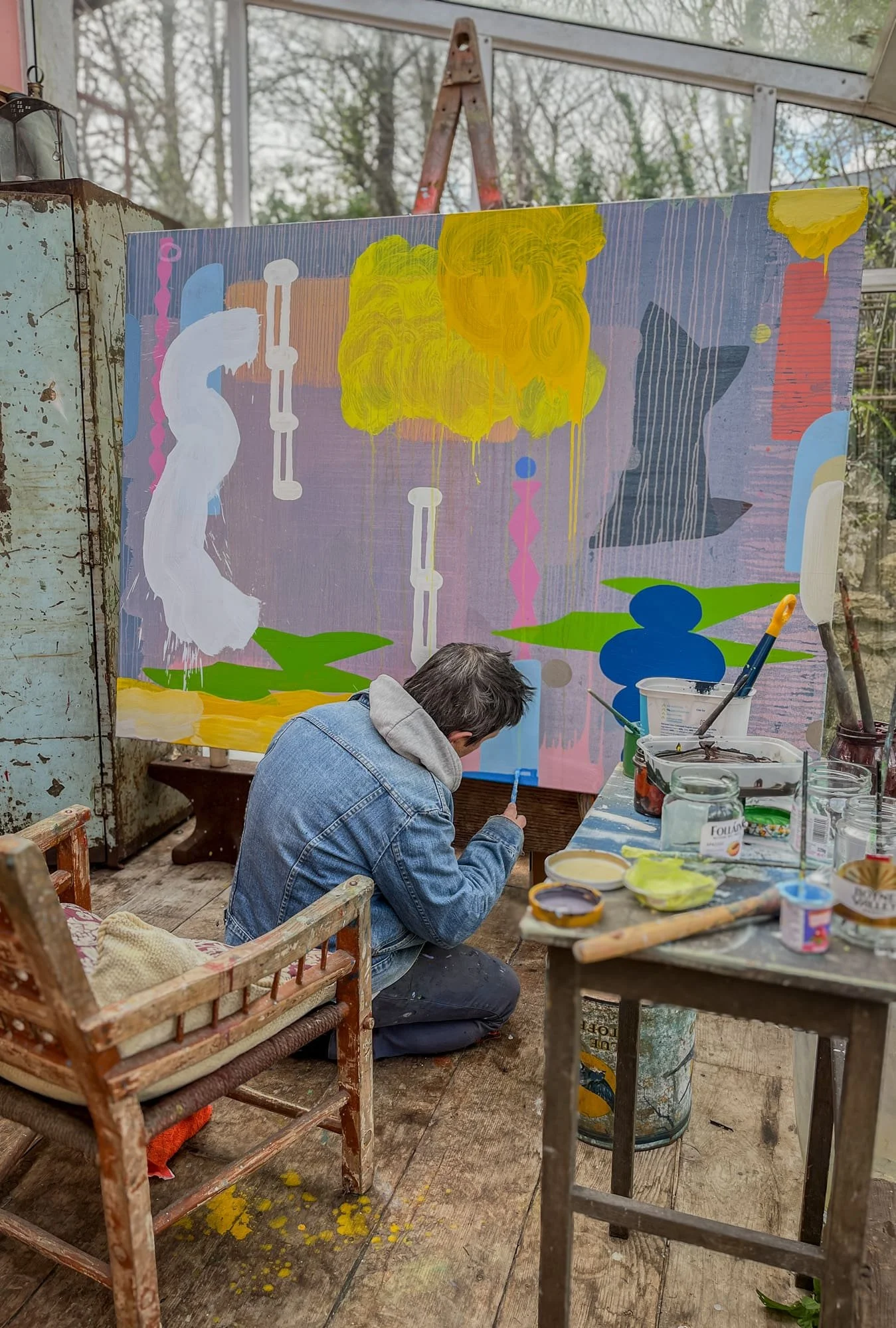 Irish artist kneeling to work on large abstract oil painting in glass studio, yellow green blue and pink forms on lavender ground, paints and jars on table beside him, Martin Finnin West Cork