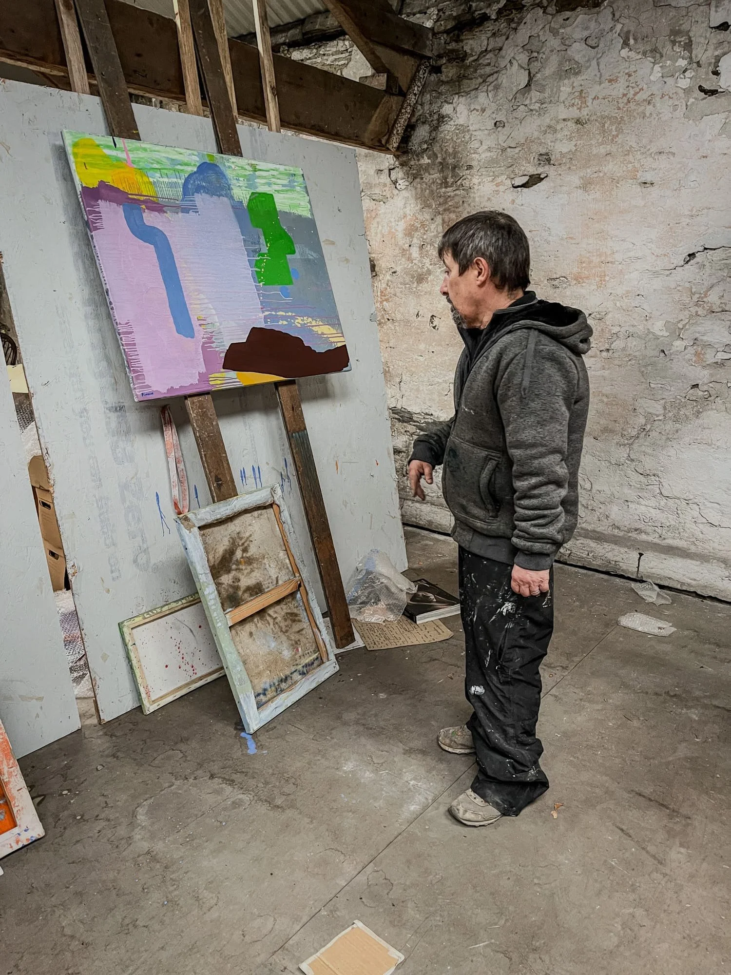Irish artist Martin Finnin standing back to look at abstract oil painting on easel in raw stone West Cork studio, lavender pink green and brown canvas, painter studying work in progress