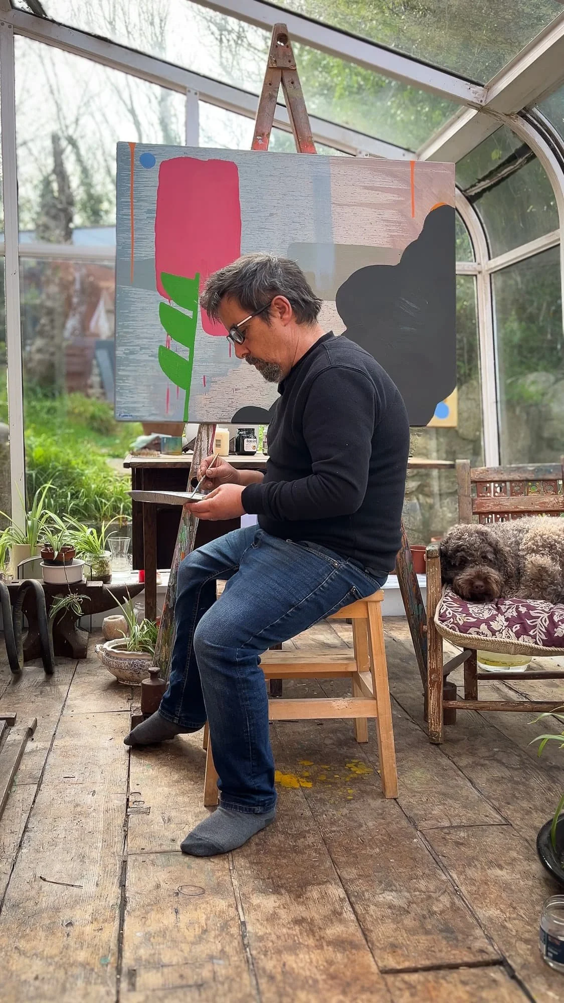 Irish artist painting in West Cork studio, abstract canvas on easel with pink red green and dark forms, dog resting in chair beside artist, Martin Finnin