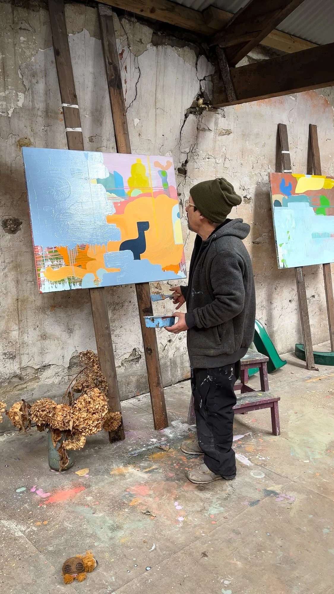 Irish artist Martin Finnin standing back to study abstract oil painting on easel, blue orange and yellow canvas nearly complete, dried hydrangeas on studio floor, two paintings on easels in West Cork studio