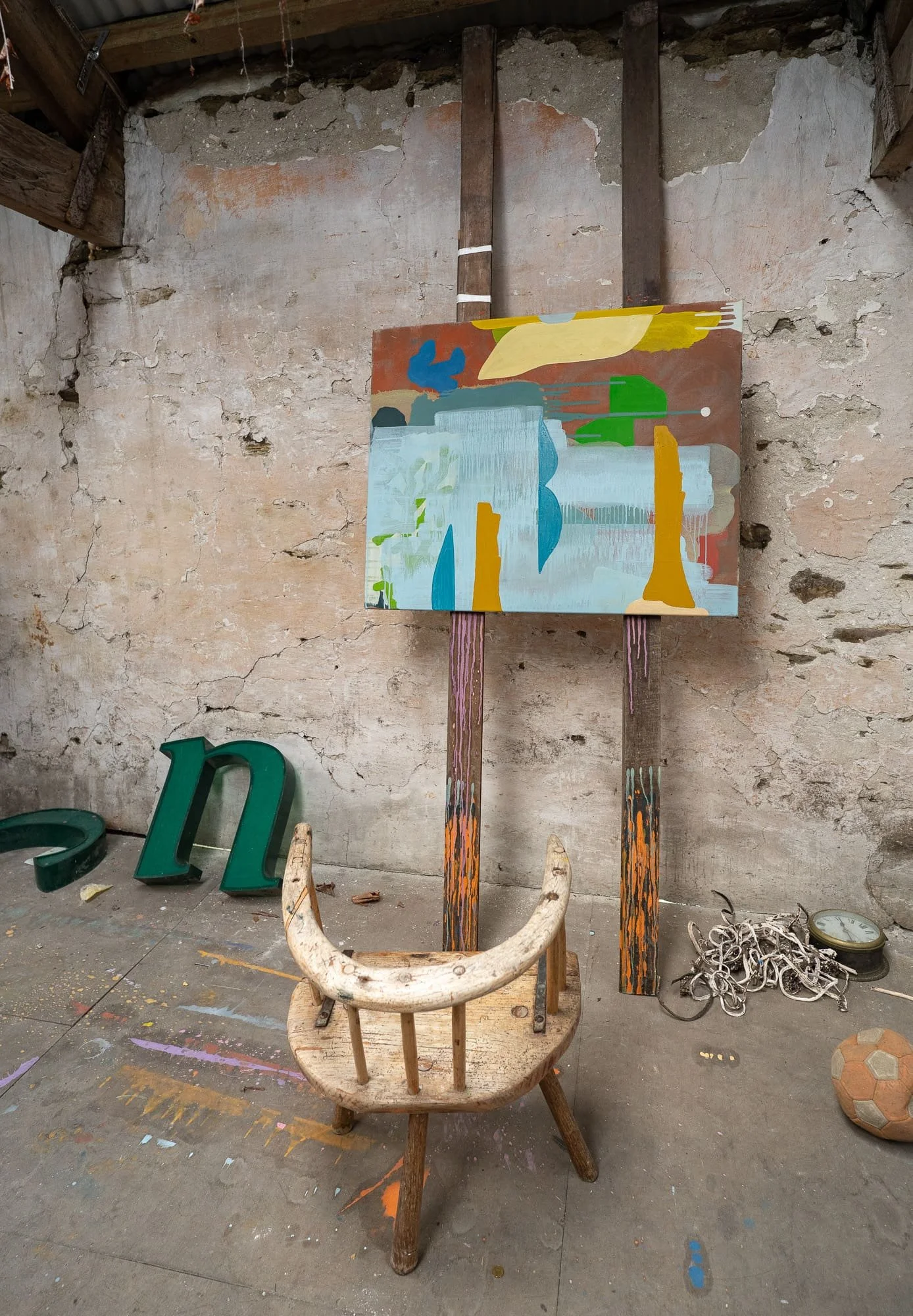 Abstract oil painting on easel in West Cork studio, terracotta yellow blue and green forms, found objects and studio debris on floor including green letters and old clock, Martin Finnin Irish artist