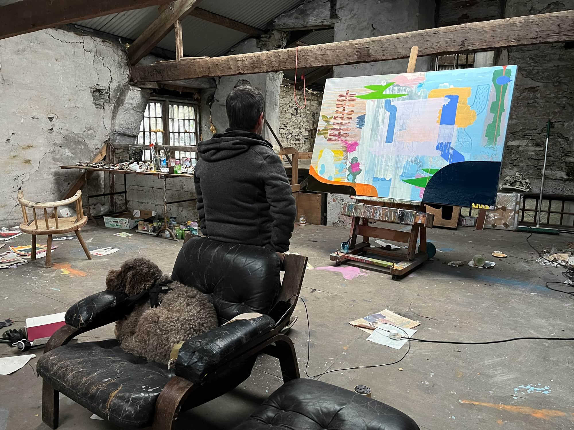 Irish artist seated in leather chair with dog studying large abstract oil painting on easel in West Cork studio, blue green orange and navy forms, stone-walled studio interior, Martin Finnin
