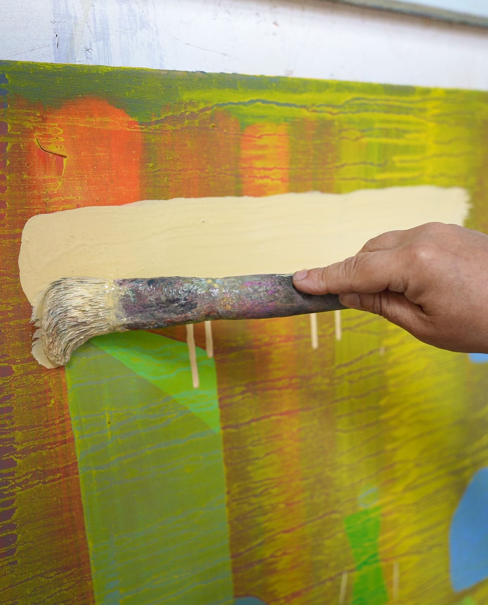 Close up of large brush applying pale wash over orange yellow and green oil paint layers, abstract painting in progress, West Cork studio, Martin Finnin Irish artist