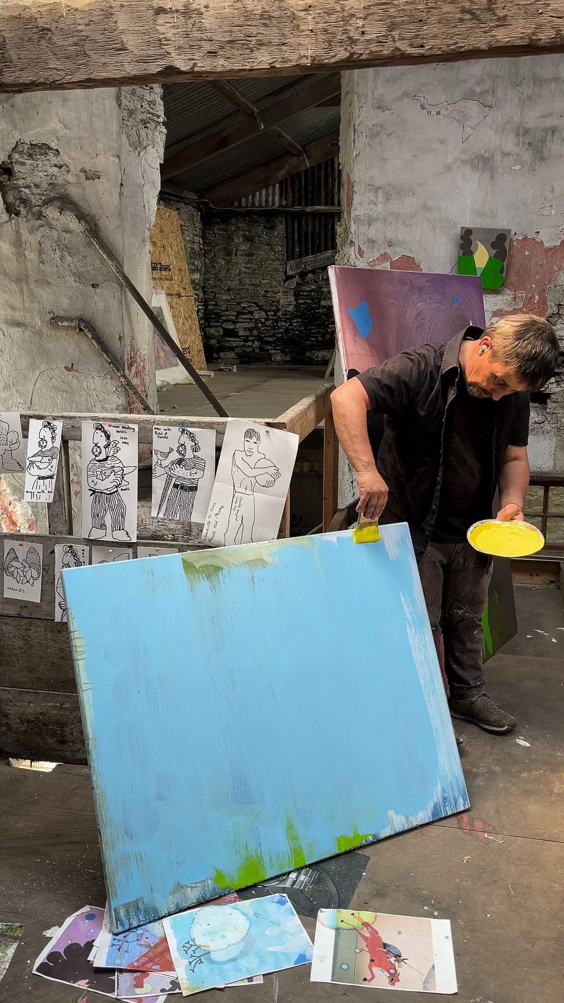 Irish artist Martin Finnin applying first yellow marks to large blue abstract canvas leaning against wall in West Cork stone studio, early stage oil painting in progress