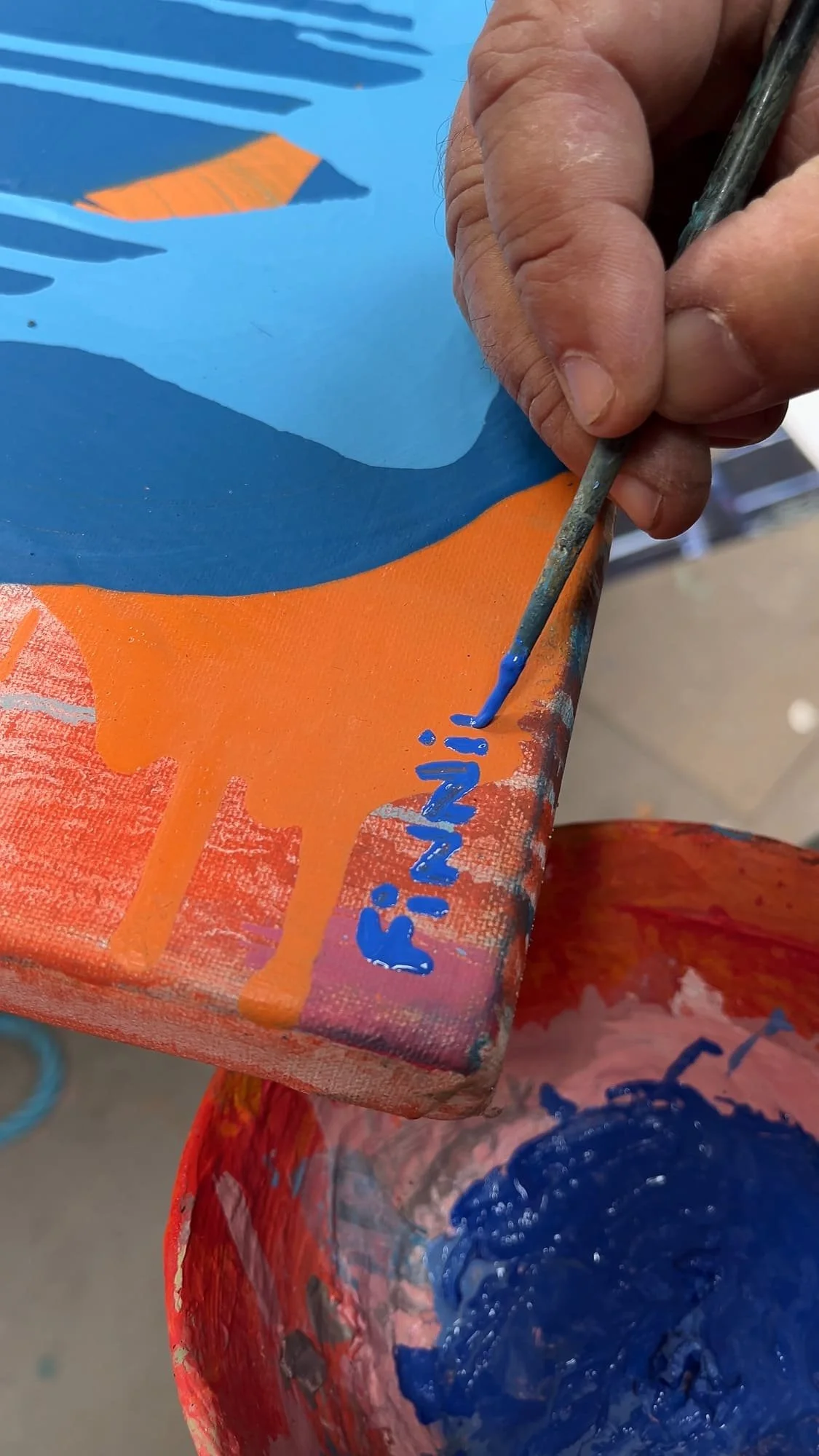 Close up of artist's hand signing abstract oil painting on canvas edge in blue paint, orange and blue canvas, Martin Finnin signing finished original painting in West Cork studio