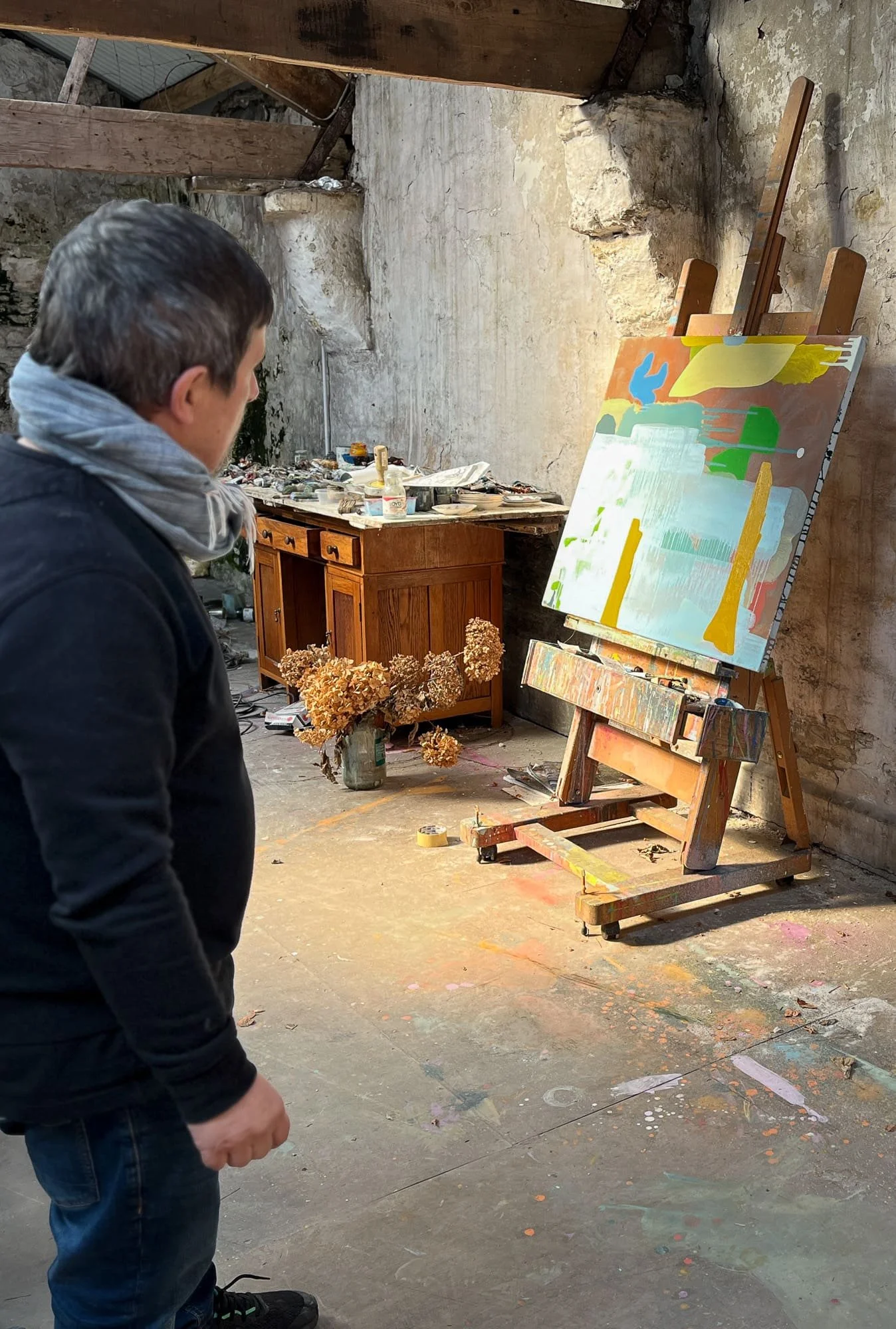 Irish artist standing back to study abstract oil painting on easel in stone-walled West Cork studio, dried hydrangeas and antique desk in background, Martin Finnin
