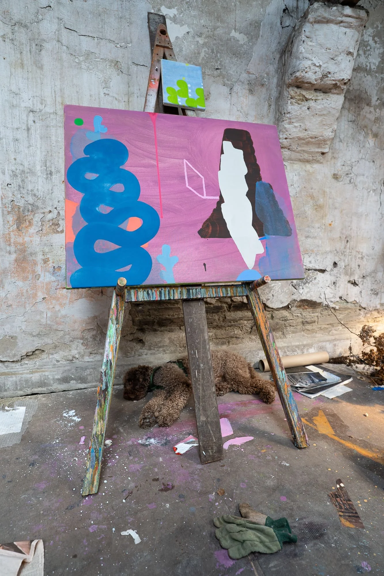 Abstract oil painting in progress on easel in West Cork studio, pink canvas with bold blue wave form developing, dog sleeping beneath easel on paint-splattered studio floor, Martin Finnin