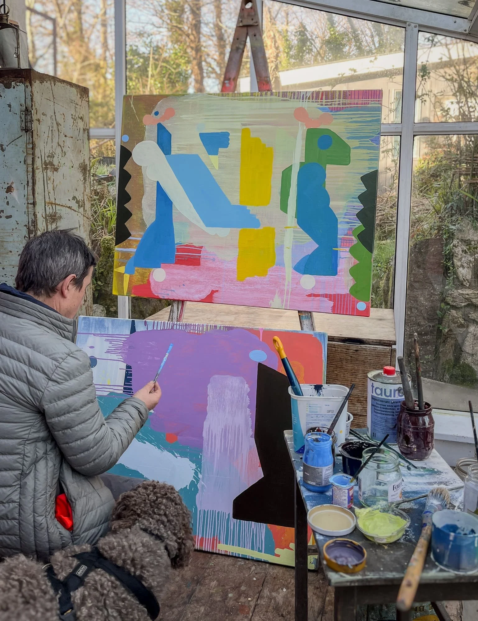 Irish artist painting in glass studio with dog beside him, finished canvas on easel behind and new work in progress below, blue yellow green and pink forms, Martin Finnin West Cork