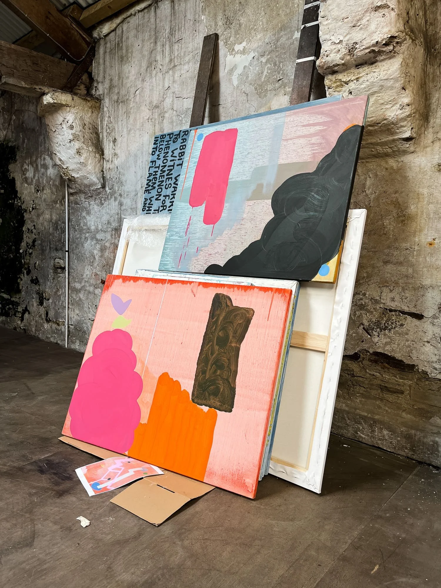 Multiple abstract oil paintings in progress leaning against raw stone wall in West Cork studio, pink and black cloud canvas above coral orange canvas, Martin Finnin